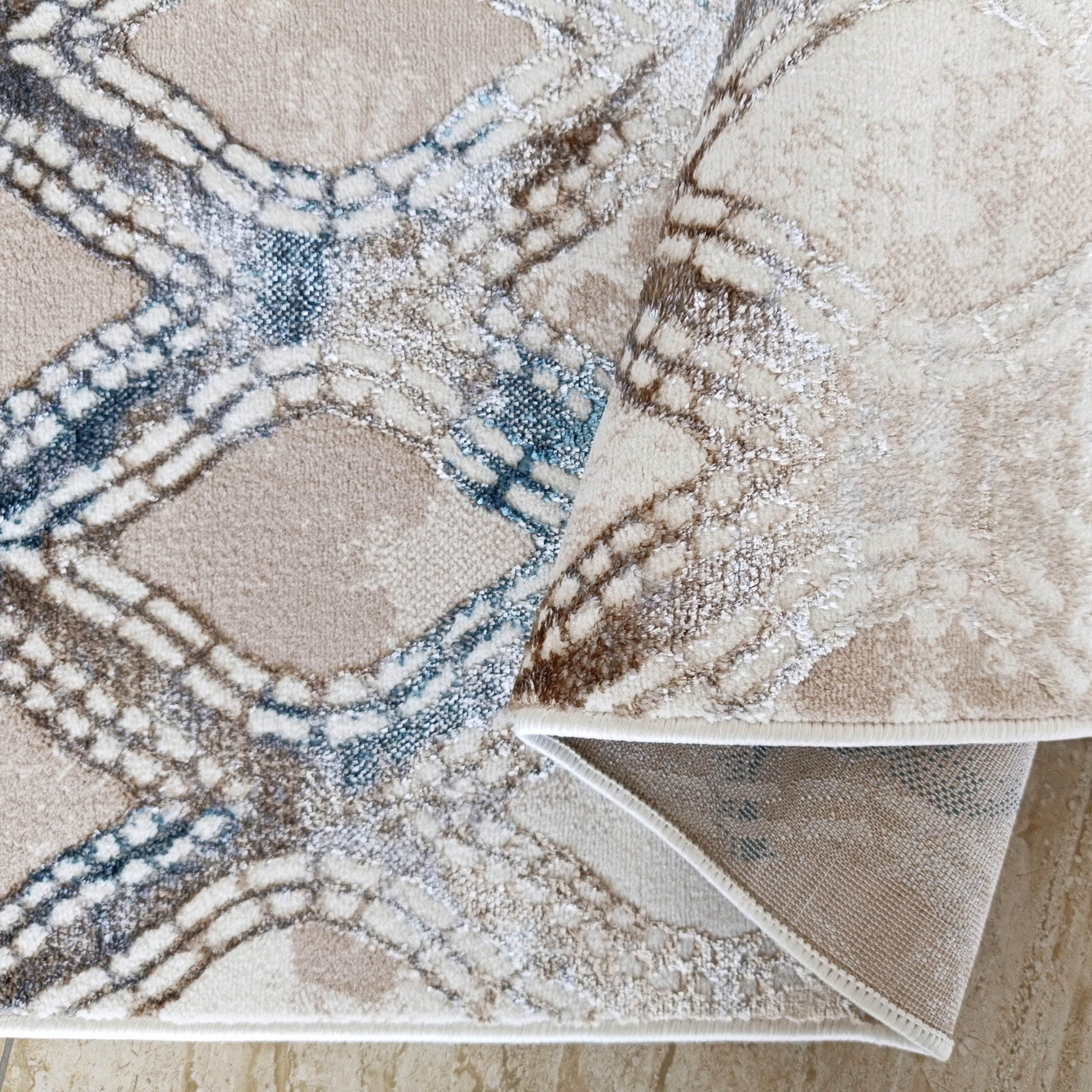 Tapis aux tons beige, crème, taupe et bleu-gris avec des motifs géométriques texturés, enroulé sur un sol carrelé clair