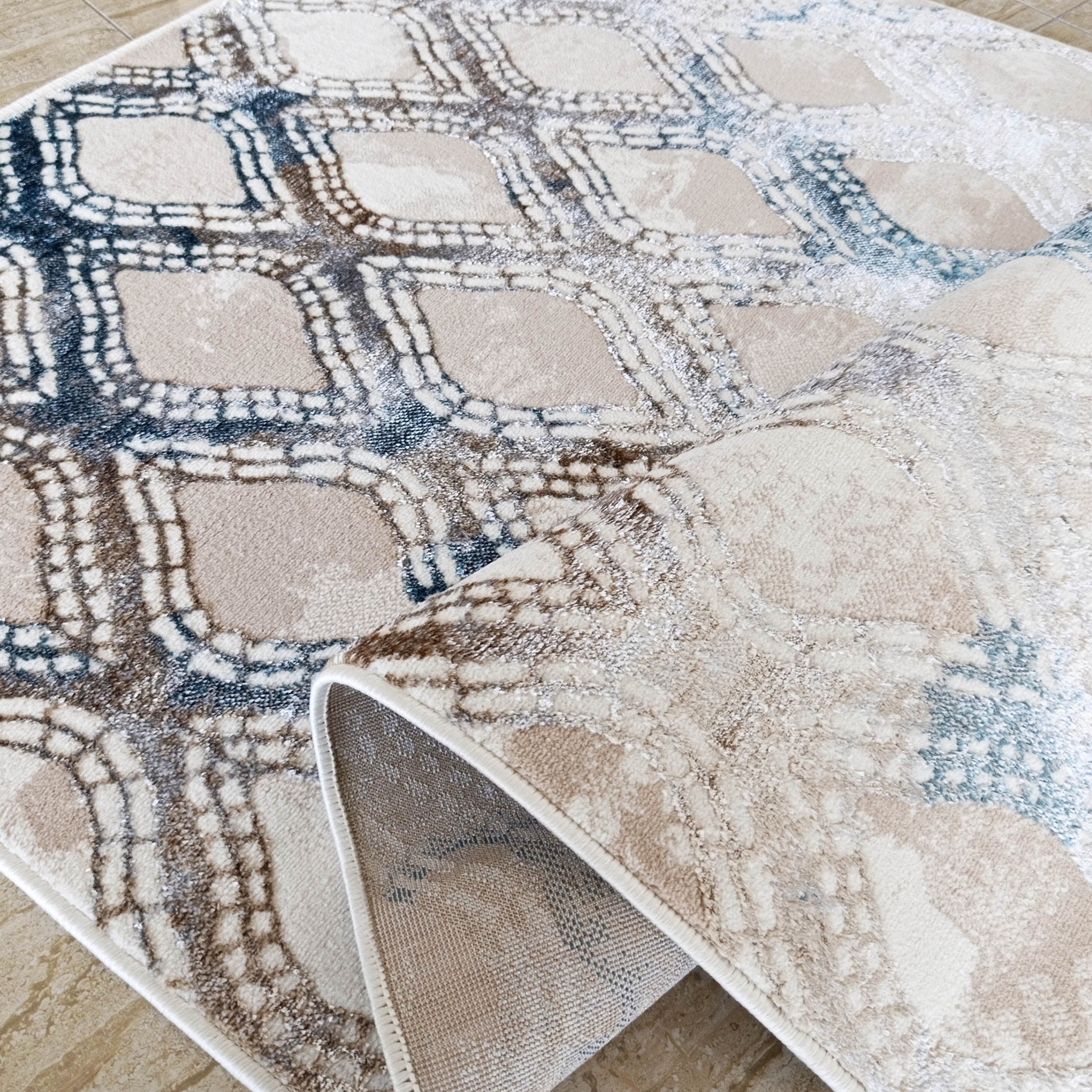 Tapis moelleux beige, bleu et marron aux motifs géométriques texturés, partiellement roulé sur un sol carrelé clair