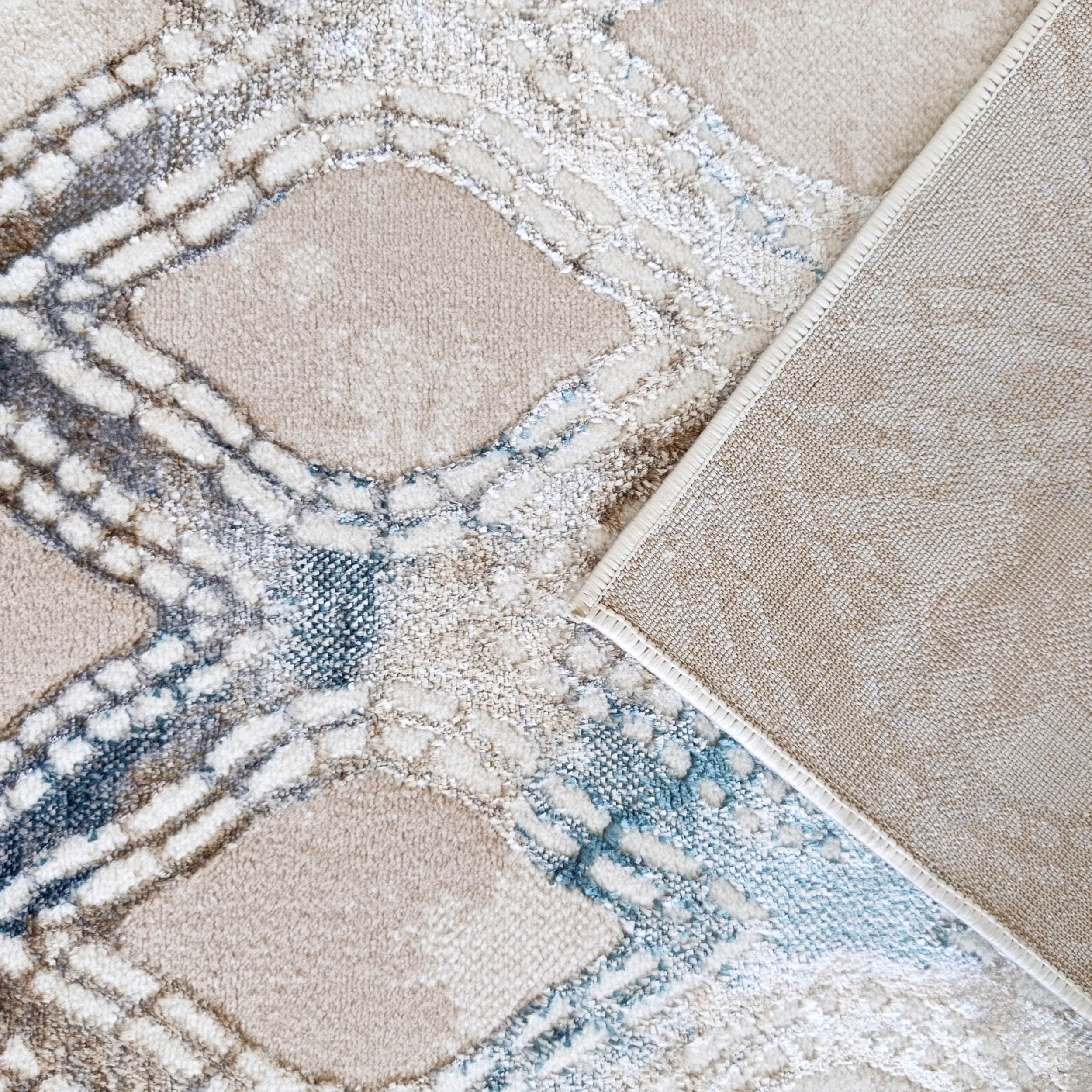 Tapis beige à losanges en lignes texturées bleues, blanches et brunes, au revers en toile beige visible sur un côté