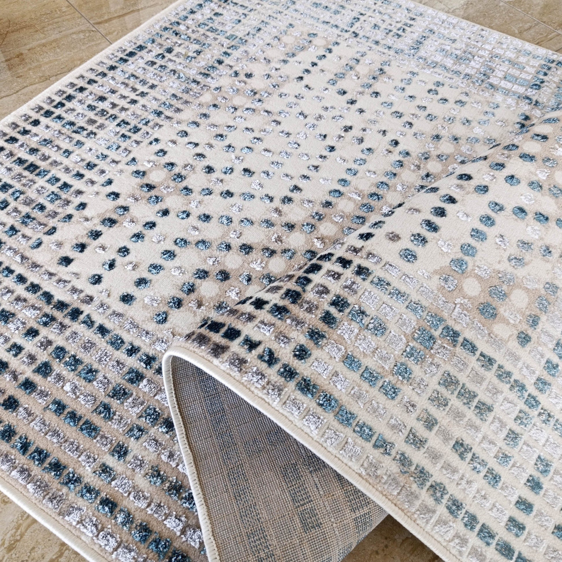 Tapis beige à motifs de carrés et de points texturés bleus et gris, partiellement enroulé sur un sol carrelé clair