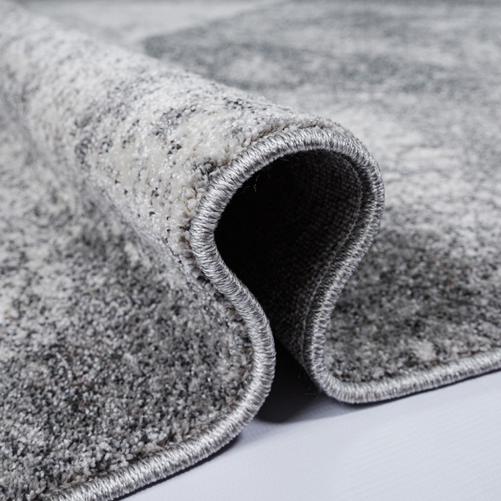 Gros plan sur un tapis texturé, partiellement roulé, montrant des nuances de gris claires et foncées avec un motif abstrait moucheté