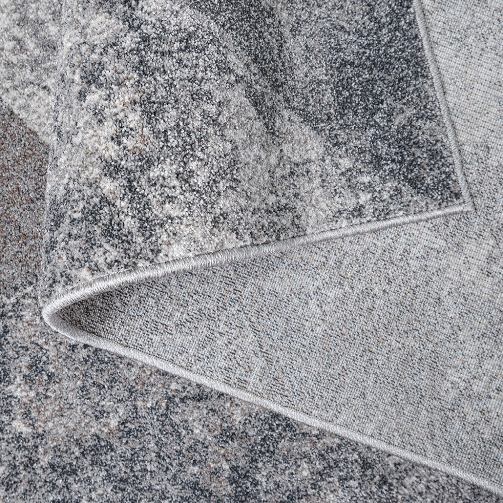 Tapis moelleux aux motifs abstraits mouchetés en nuances de gris, blanc et beige. Un coin est relevé, montrant son envers tissé et son bord fini