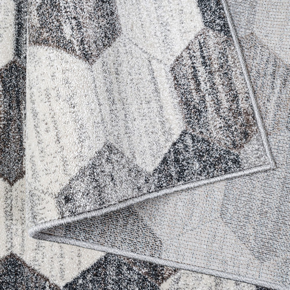 Tapis à surface texturée aux motifs géométriques entrelacés en teintes de gris, crème et brun. Un coin est replié, révélant la sous-face tissée