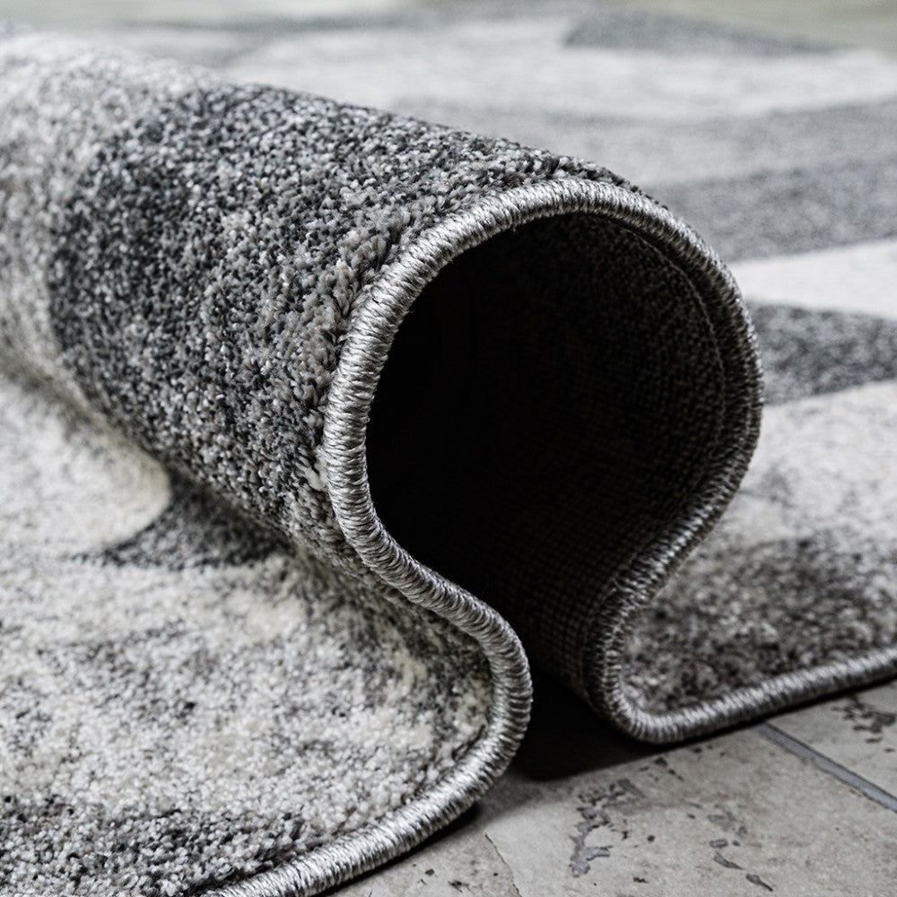 Tapis gris et blanc à motifs, partiellement roulé sur un sol carrelé, dévoilant sa texture douce et moelleuse