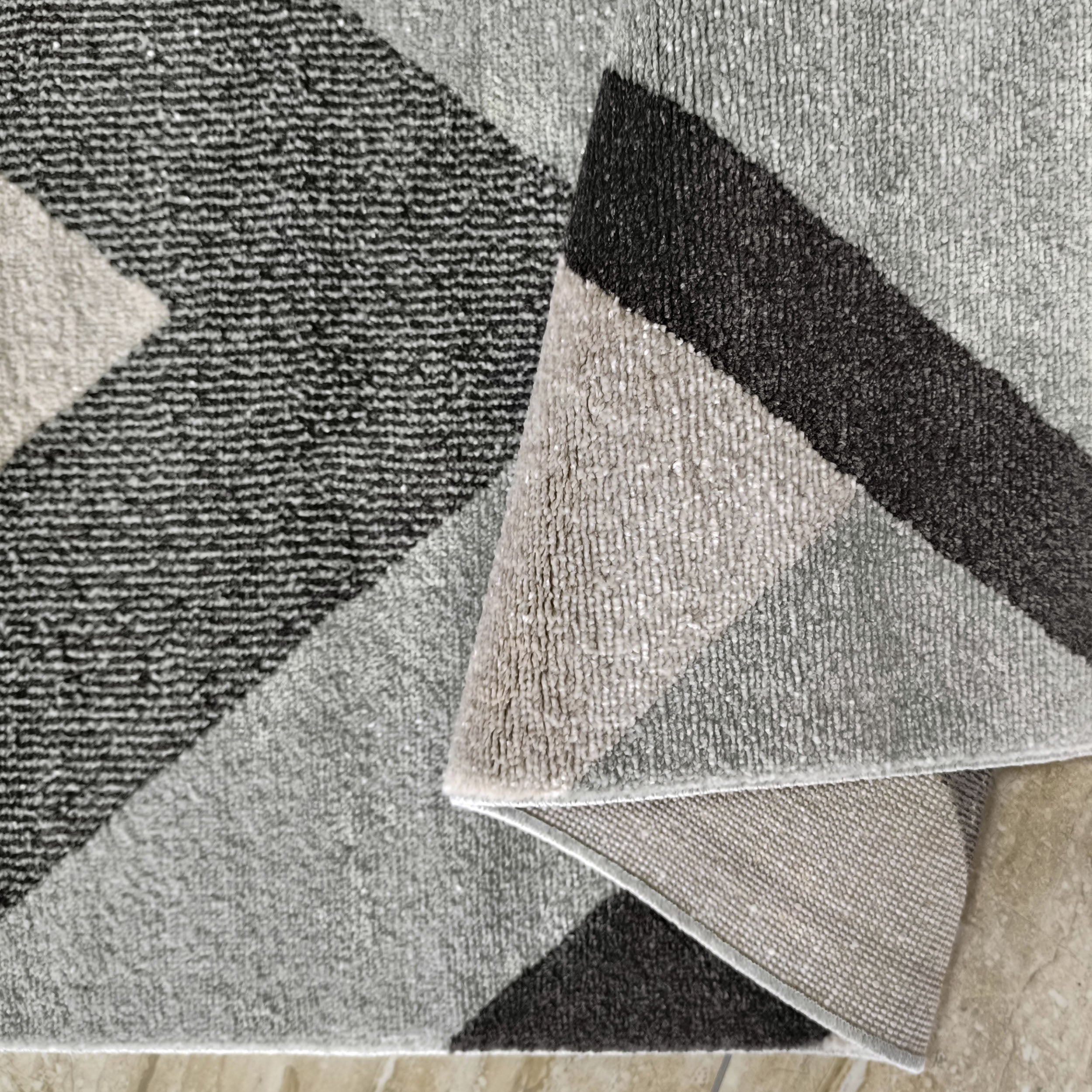Tapis textile à la texture douce, orné de motifs géométriques en blocs gris foncé, gris moyen et crème, partiellement plié