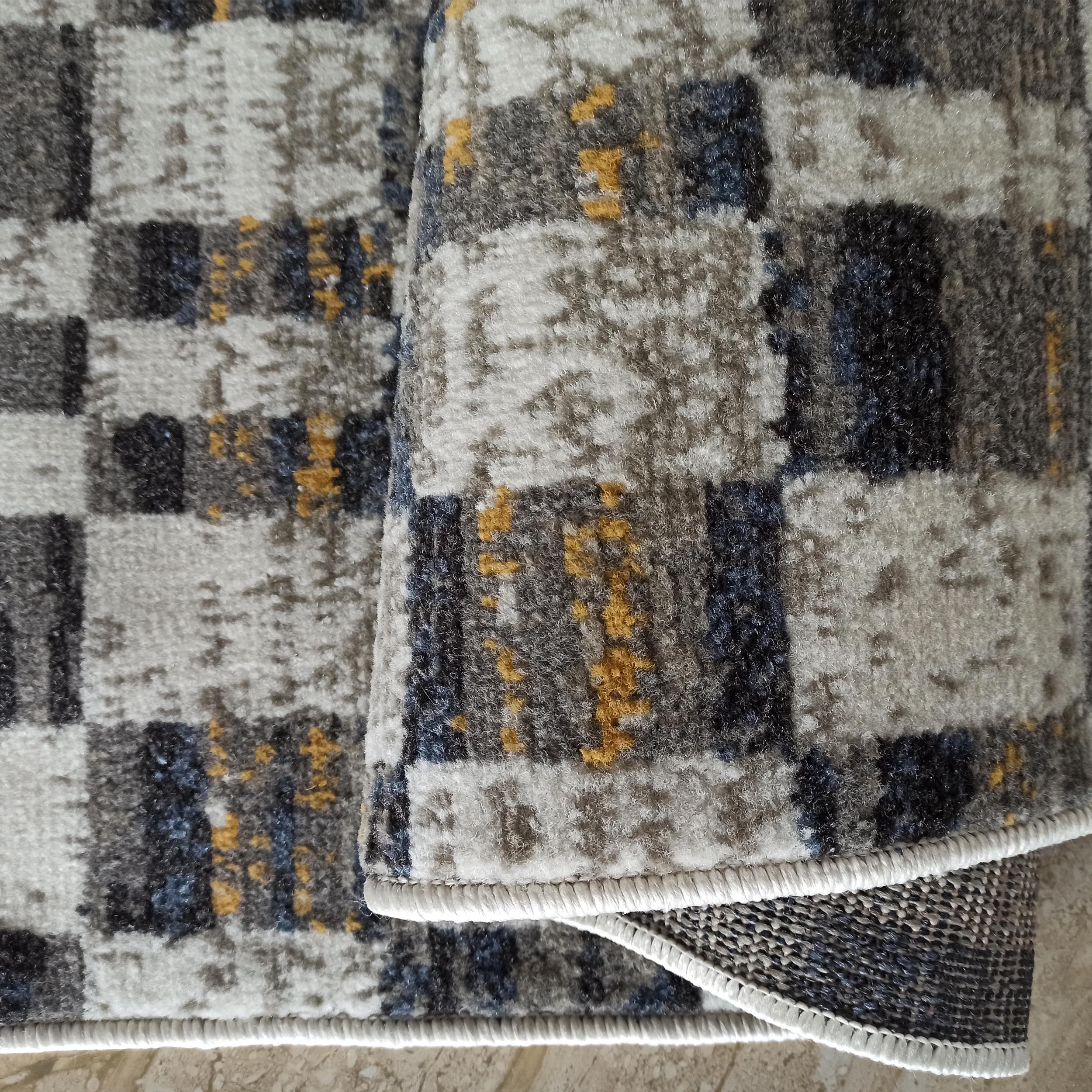 Tapis texturé à motifs abstraits gris, blanc, bleu foncé et jaune, une partie est roulée révélant le dessous tissé