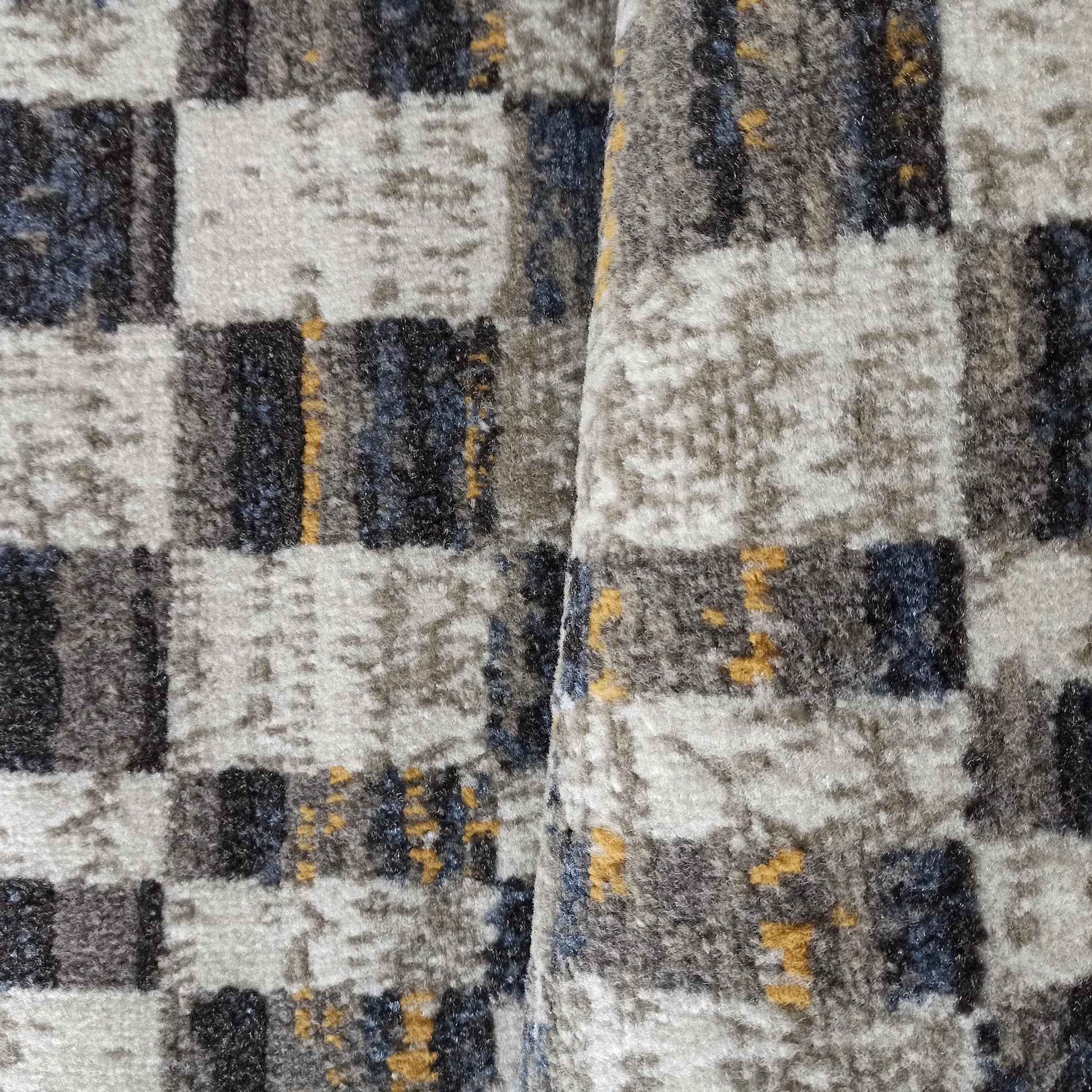 Tapis à texture douce et dense, avec un motif abstrait de carreaux mêlant des nuances de gris, noir, bleu et jaune