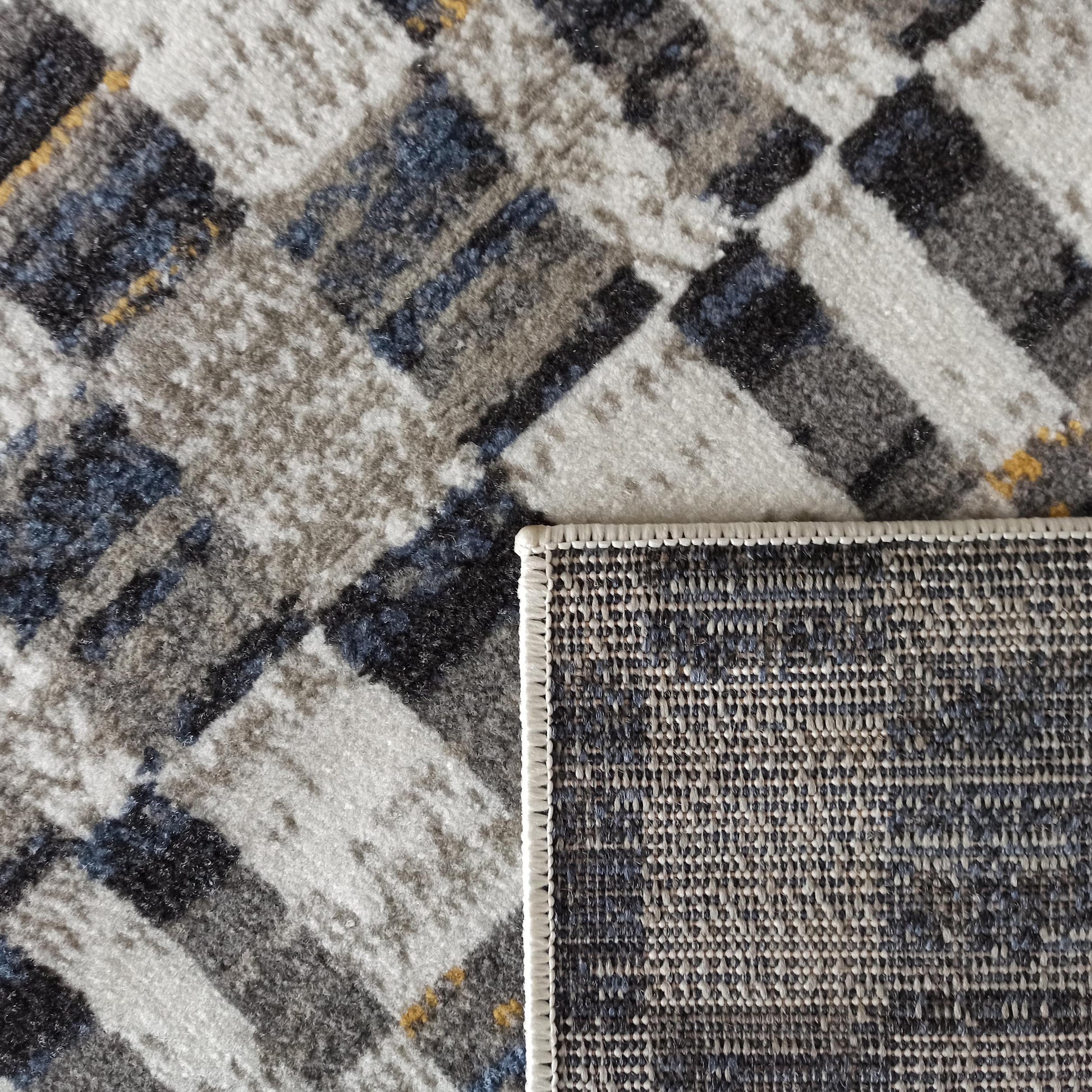 Tapis à motifs géométriques abstraits gris, blanc, bleu et noir, avec des touches jaunes, montrant sa texture douce et son envers tissé