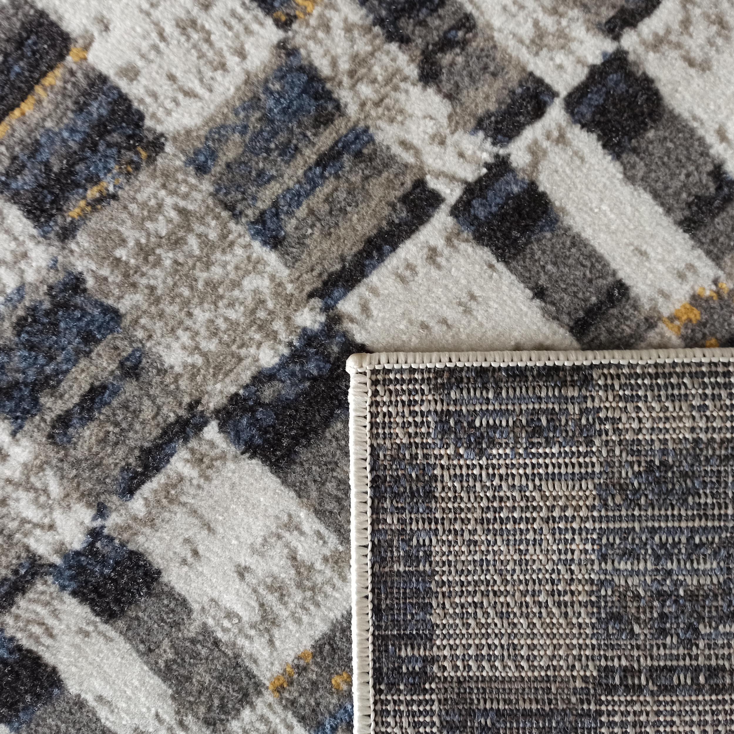 Tapis à motifs géométriques abstraits gris, blanc, bleu et noir, avec des touches jaunes, montrant sa texture douce et son envers tissé