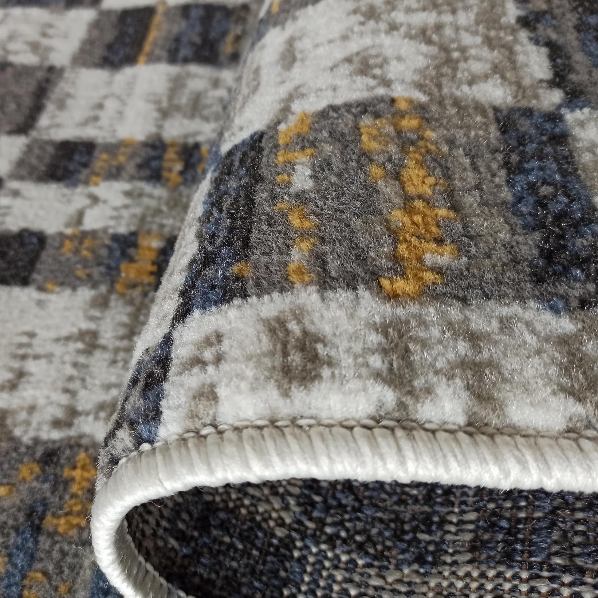 Gros plan d'un tapis à texture douce, présentant des motifs abstraits dans des teintes de gris, blanc, bleu et jaune doré, avec son bord roulé
