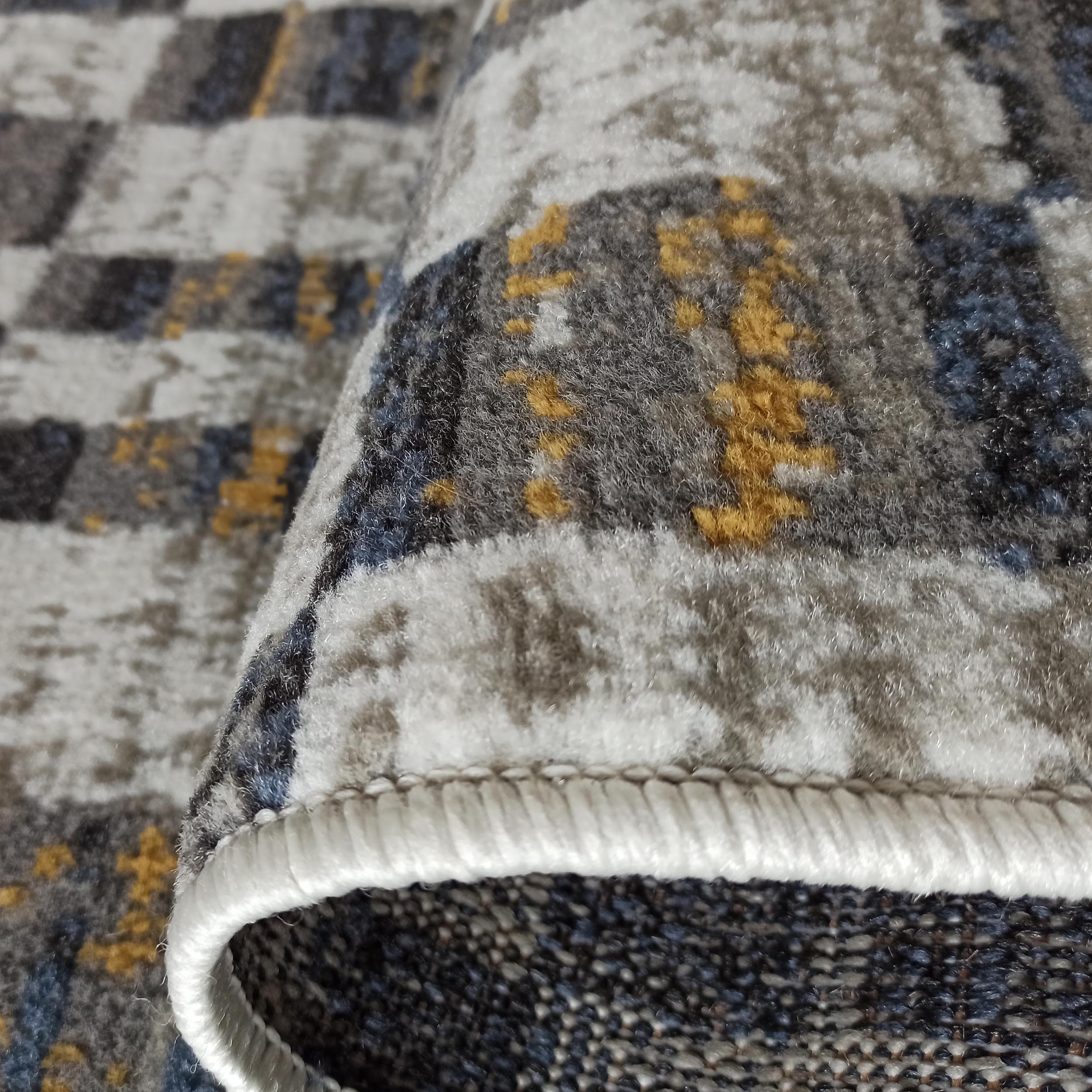 Gros plan d'un tapis à texture douce, présentant des motifs abstraits dans des teintes de gris, blanc, bleu et jaune doré, avec son bord roulé