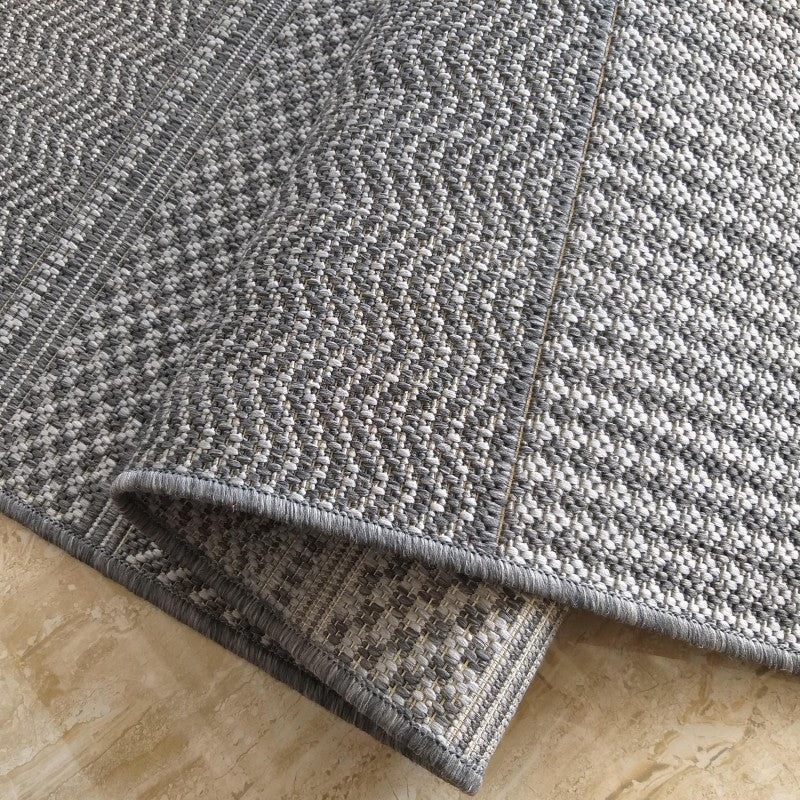 Tapis en textile tissé gris et blanc, avec un motif géométrique régulier, partiellement roulé sur un sol clair