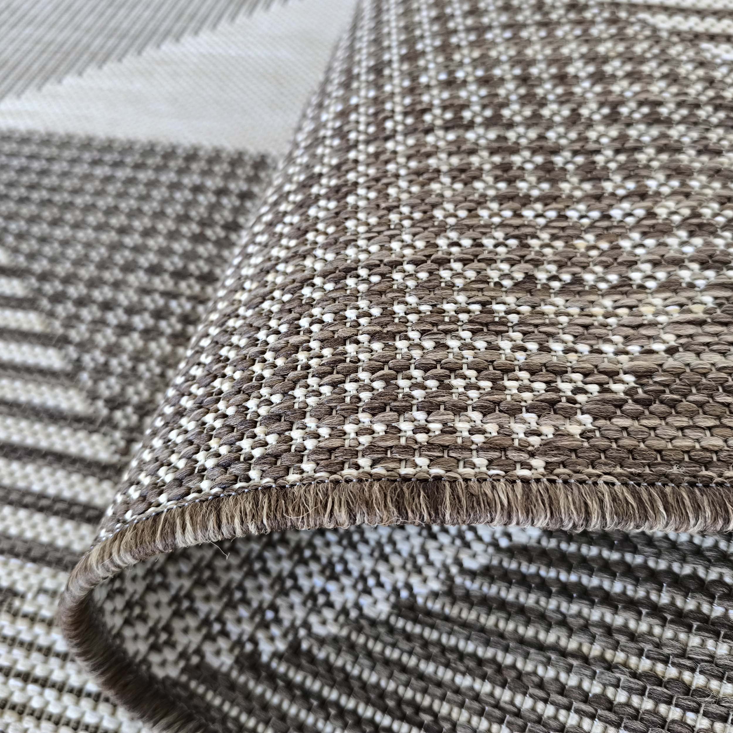 Tapis tissé en fibres apparentes, formant des motifs géométriques marron et écru, avec une bordure frangée, partiellement roulé