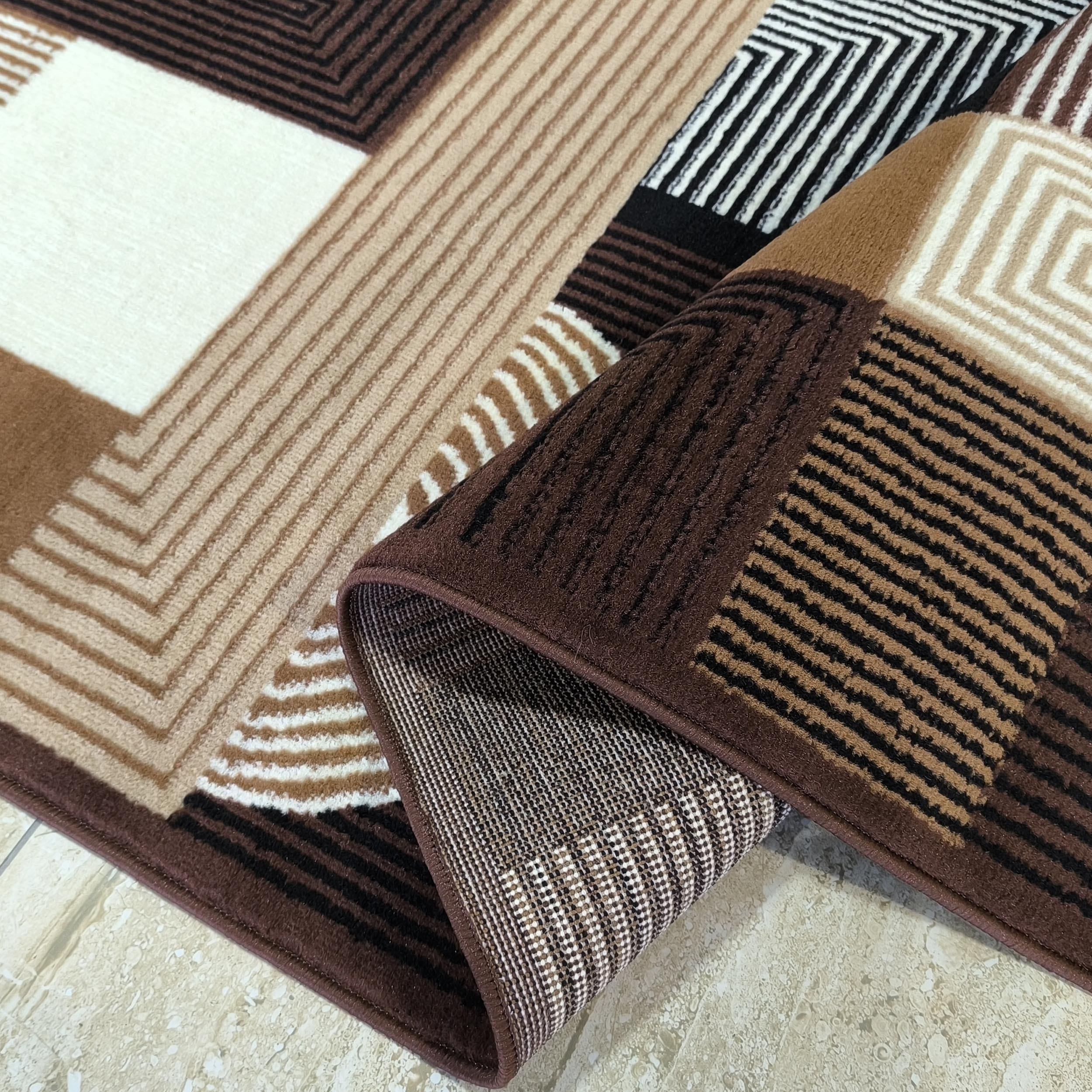 Tapis à poils courts affichant des motifs géométriques de rayures et blocs marron, beige, blanc et noir, avec une partie enroulée révélant son revers tissé