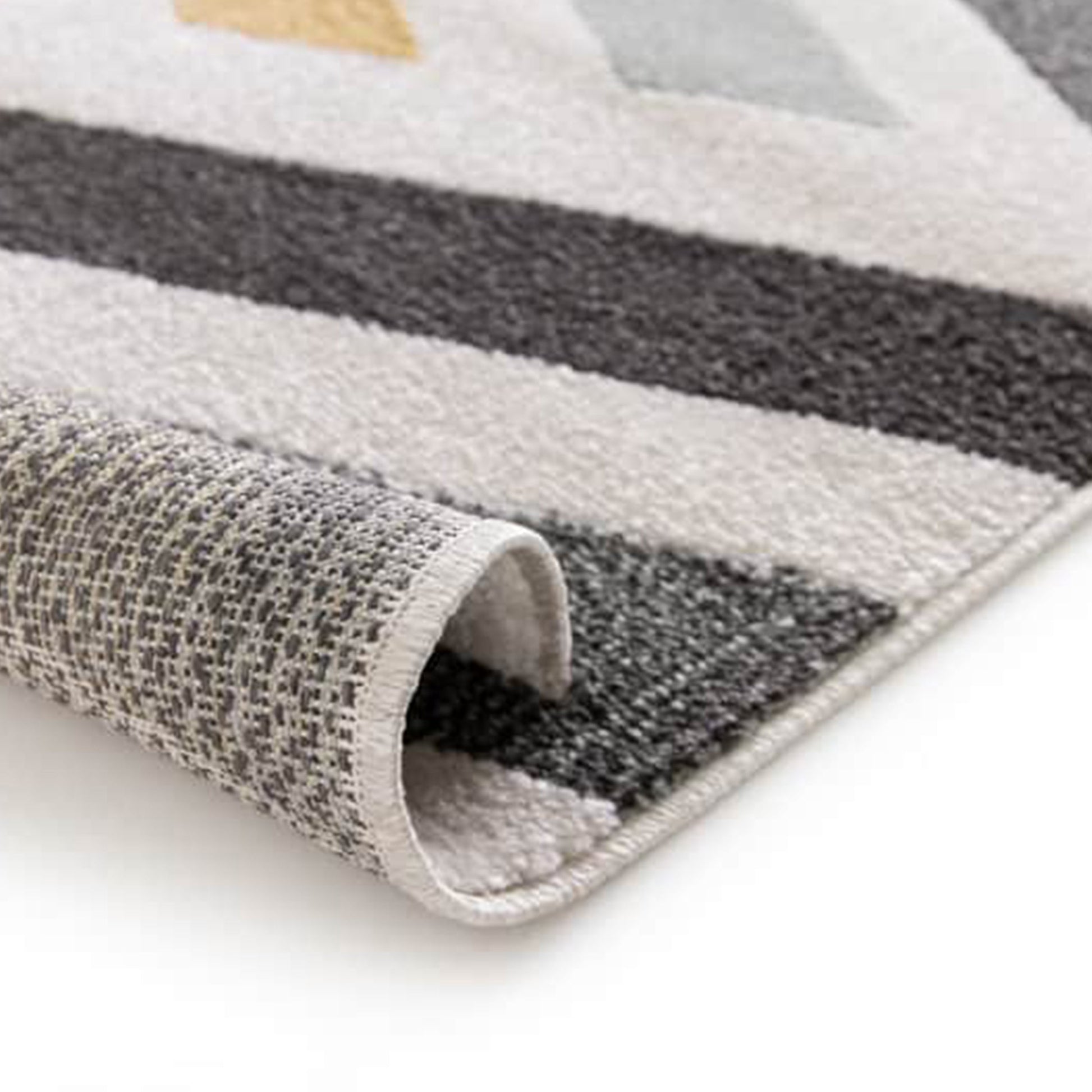 Tapis texturé à motifs géométriques de rayures diagonales gris foncé, blanc cassé et gris clair, avec un triangle jaune. Il est partiellement roulé, dévoilant son envers tissé