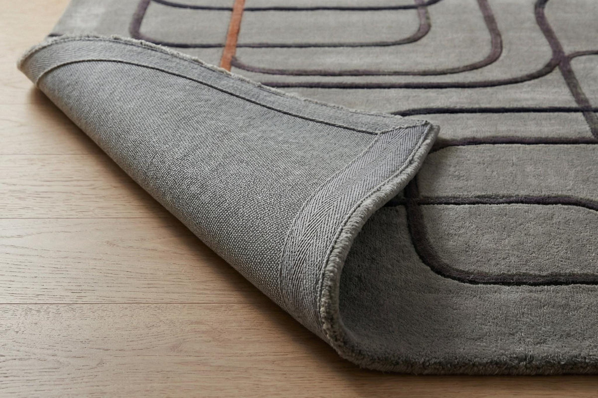 Coin replié d'un tapis gris en laine et viscose à lignes anthracite en relief, montrant son envers tissé sur un parquet