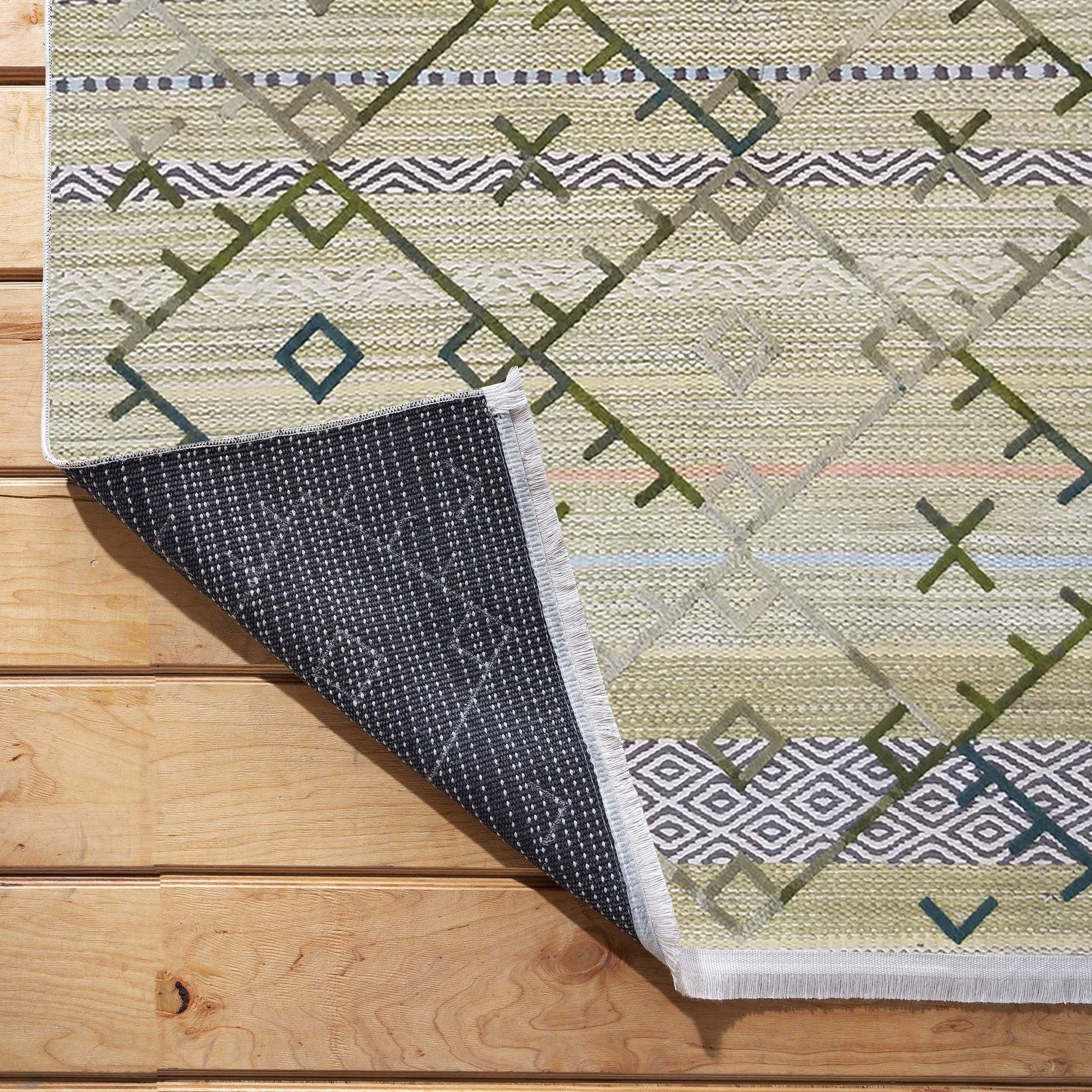 Tapis tissé à motifs géométriques verts, gris et beige clair posé sur un plancher en bois, avec un coin relevé dévoilant son envers sombre à picots