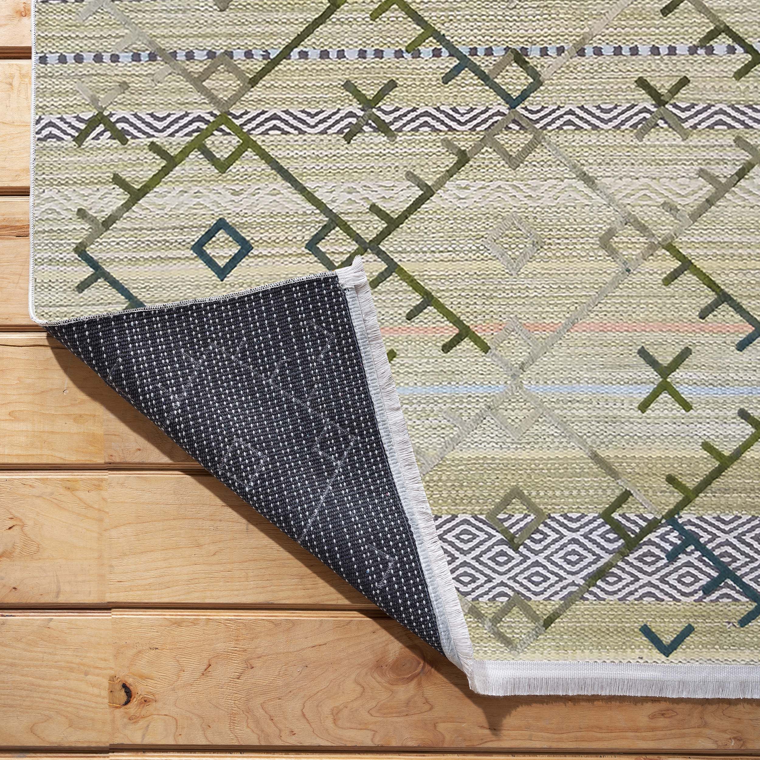 Tapis tissé à motifs géométriques verts, gris et beige clair posé sur un plancher en bois, avec un coin relevé dévoilant son envers sombre à picots
