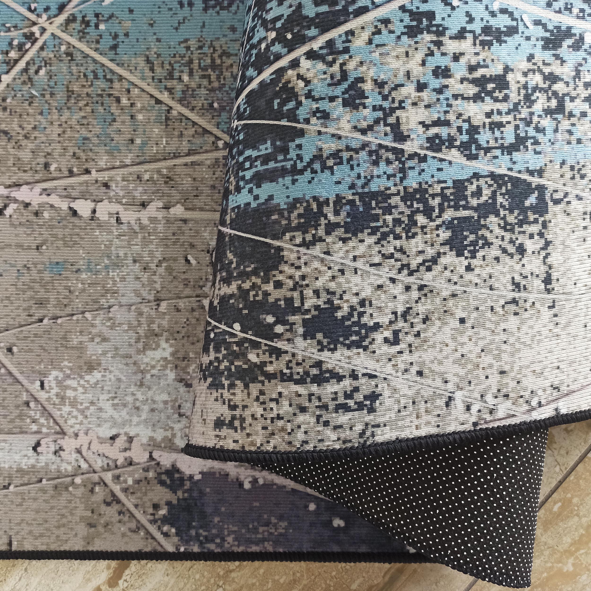 Tapis aux motifs abstraits bleus, beiges et blancs, avec des lignes claires. Sa bordure foncée et son envers noir à pois blancs sont visibles
