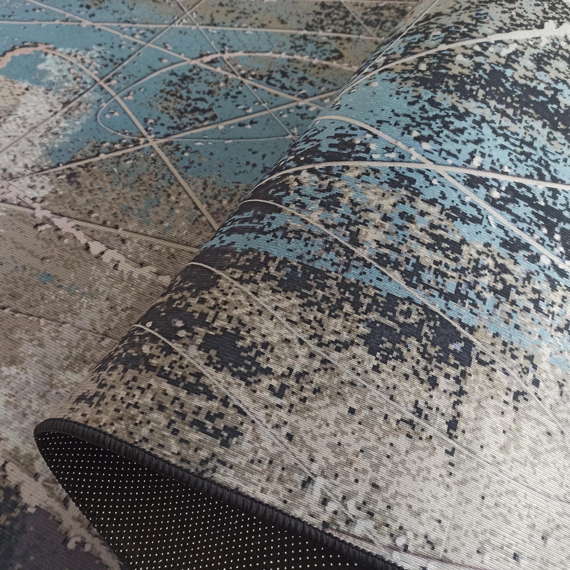 Tapis partiellement roulé aux motifs abstraits bleus, gris et noirs révélant son envers antidérapant noir à pois blancs