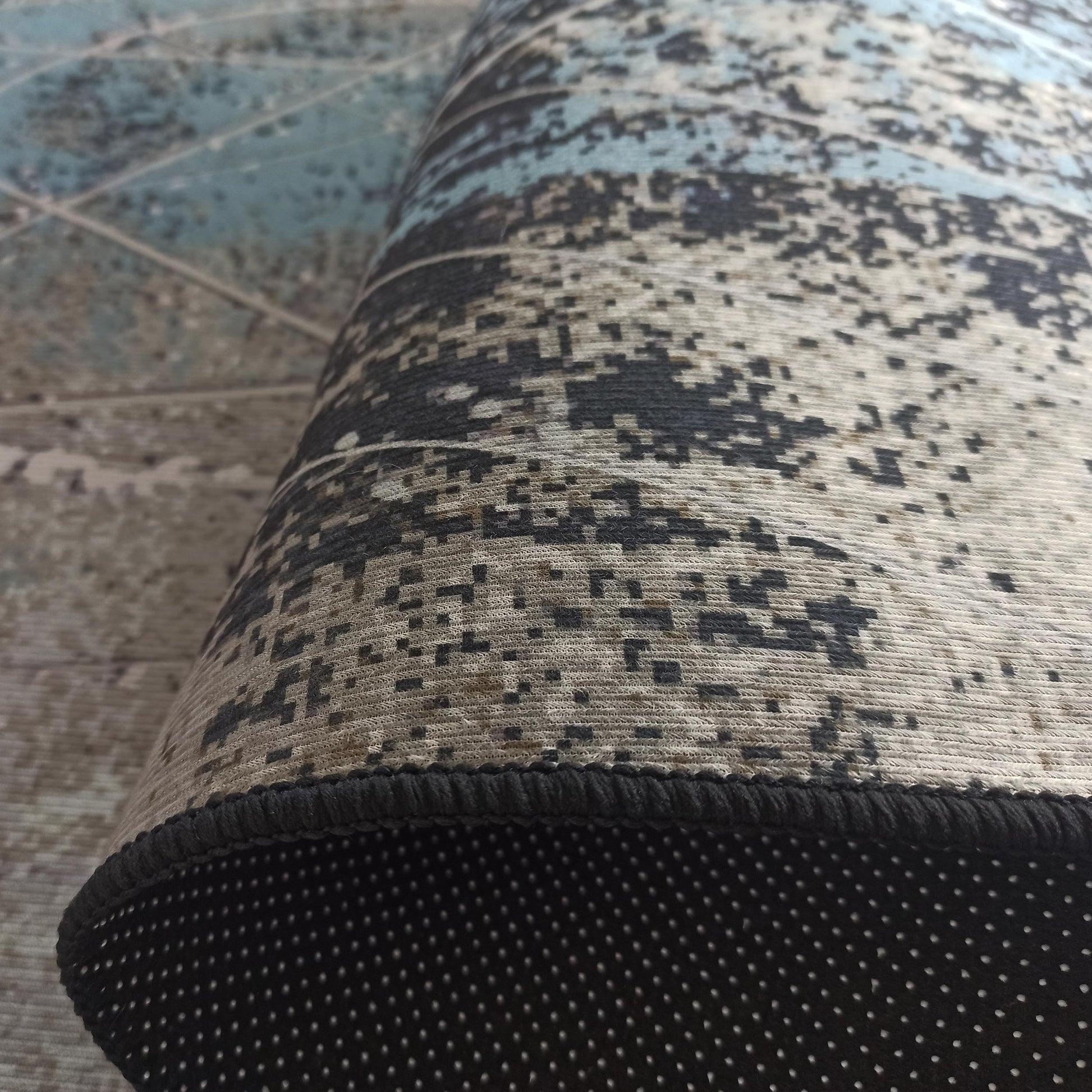 Tapis roulé avec motif abstrait pixelisé bleu, gris et beige. Le bord est noir, et le dessous antidérapant est noir à pois blancs