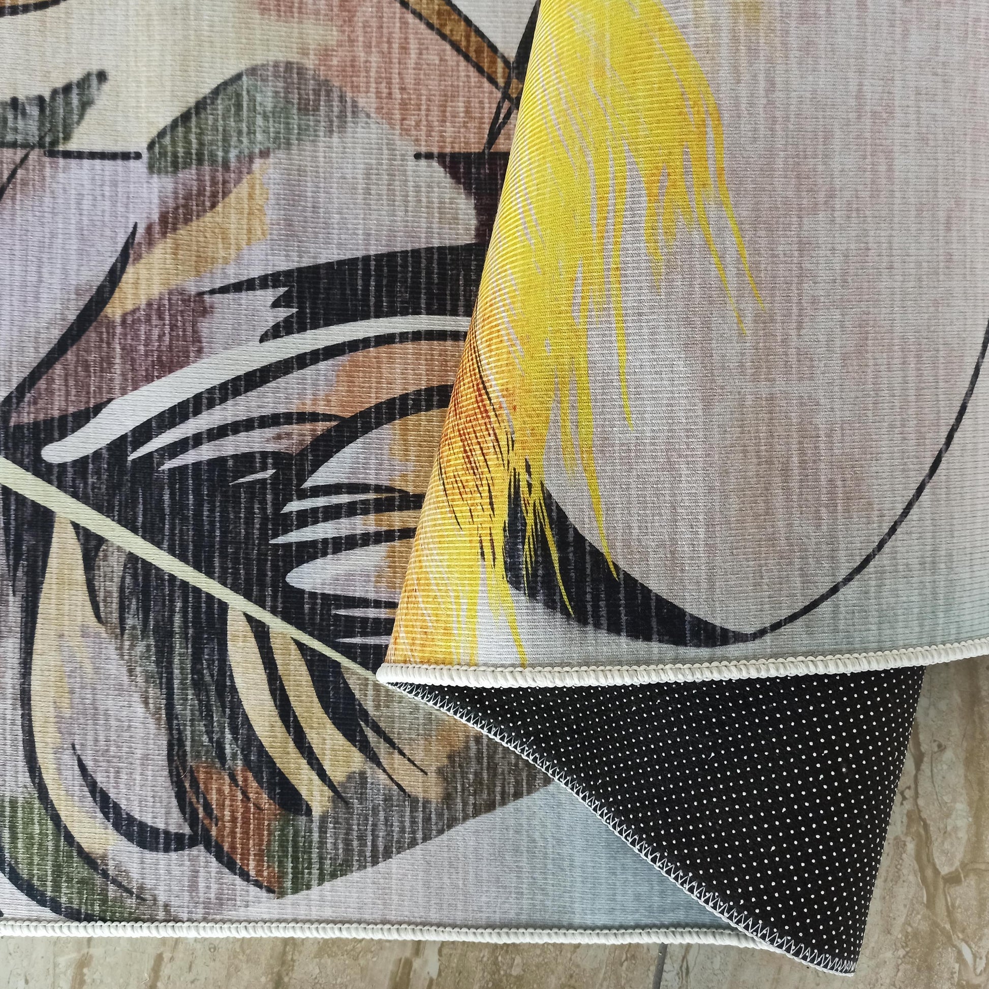 Tapis plié, motifs feuilles abstraites jaunes, noires, marron, vertes sur fond beige strié, bordure blanche, dos noir à pois blancs