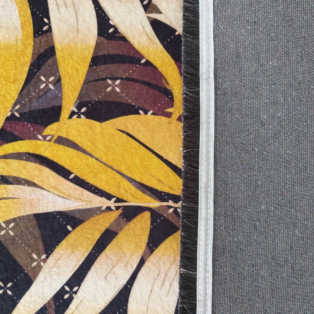Tapis doux aux motifs de feuilles stylisées jaunes et beiges sur fond sombre, avec un revers gris uni bordé d'une frange foncée et d'une couture blanche