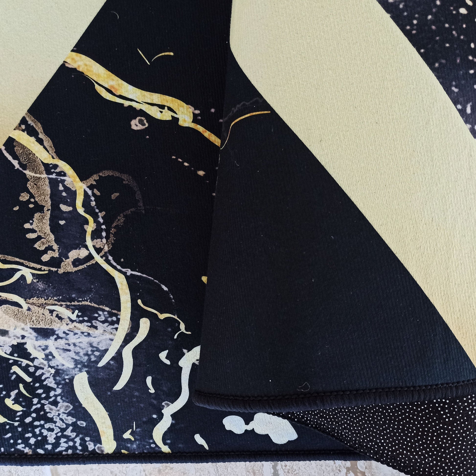 Tapis textile bicolore noir et jaune crème. Le côté noir présente des motifs abstraits dorés et blancs. Un coin soulevé montre le dos antidérapant à pois