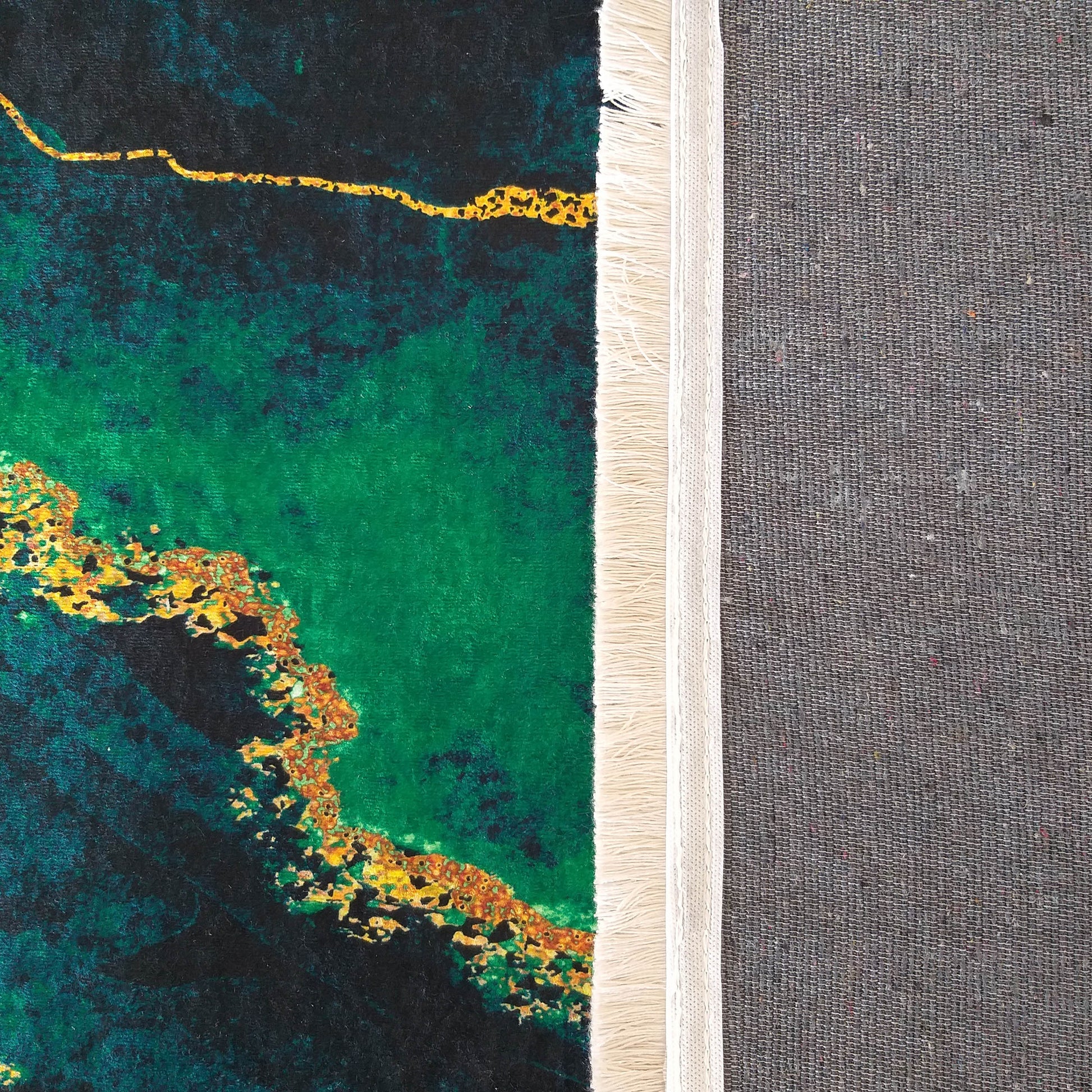 Tapis au motif abstrait vert émeraude et noir profond avec des veines et taches dorées. Une bordure frangée crème et l'envers tissé gris sont visibles