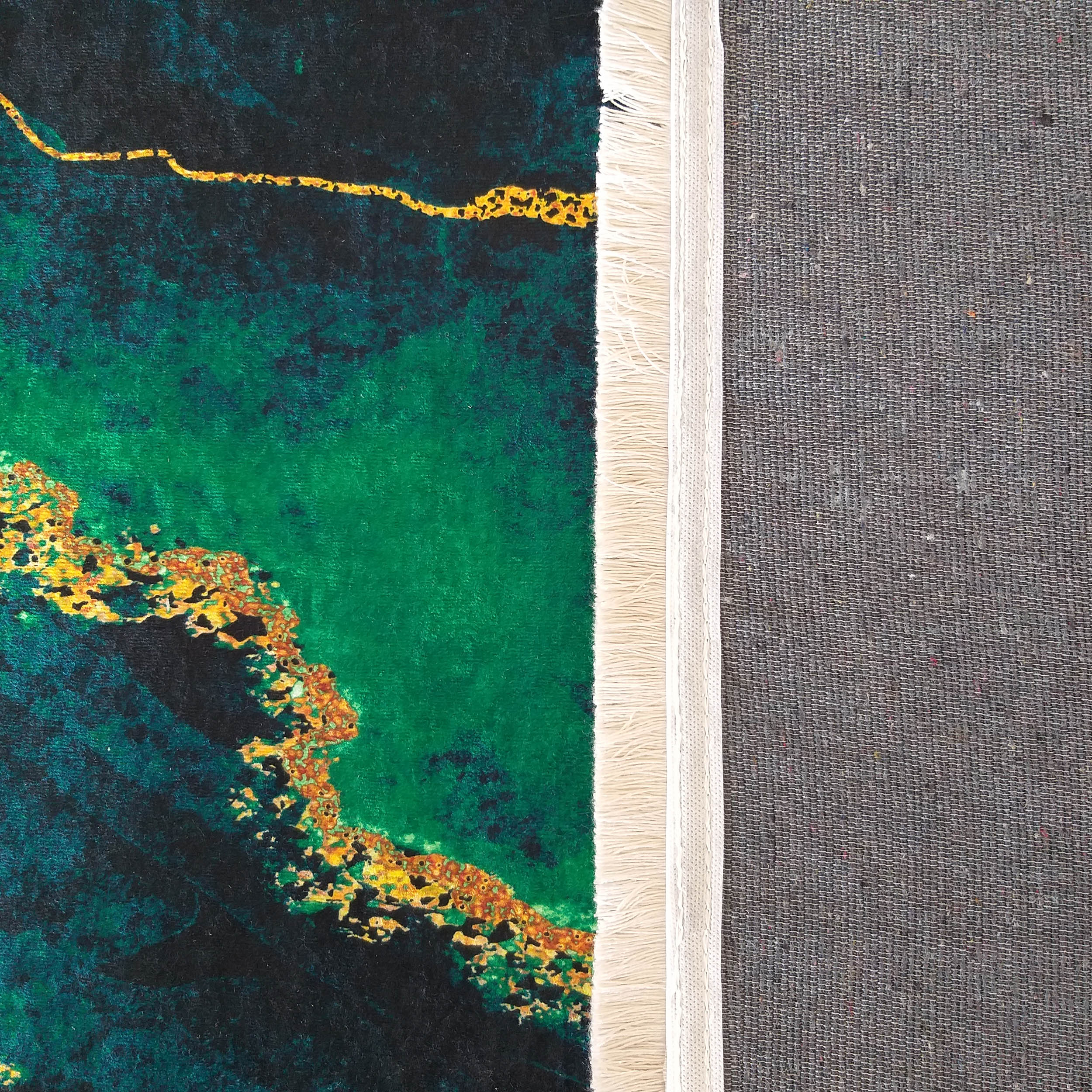 Tapis au motif abstrait vert émeraude et noir profond avec des veines et taches dorées. Une bordure frangée crème et l'envers tissé gris sont visibles