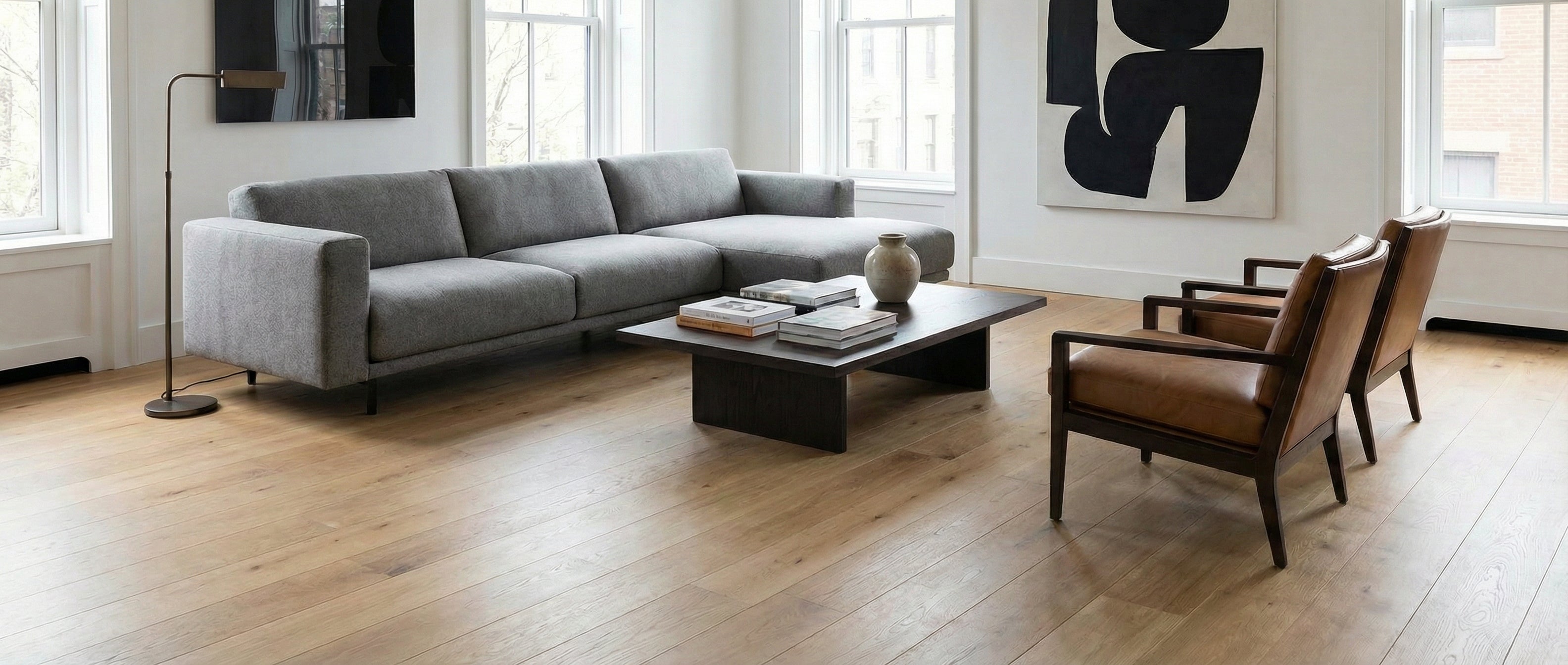 Pièce lumineuse avec canapé gris, table basse sombre, chaises cuir. Sol bois clair. Espace vide, potentiel déco