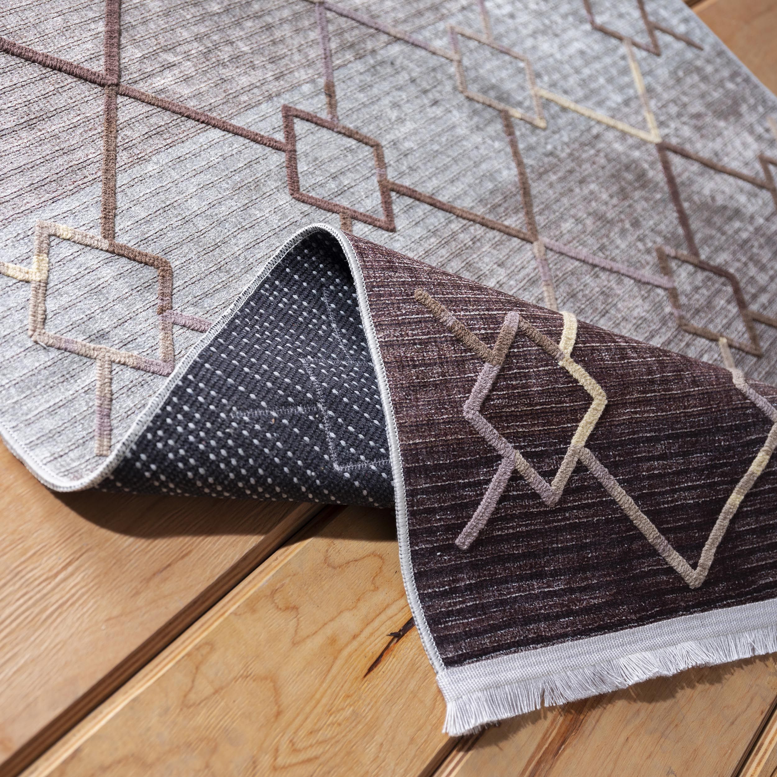 Tapis texturé gris-brun avec motifs géométriques beiges et bruns, partiellement roulé sur un plancher en bois clair, montrant sa frange et son envers à pois
