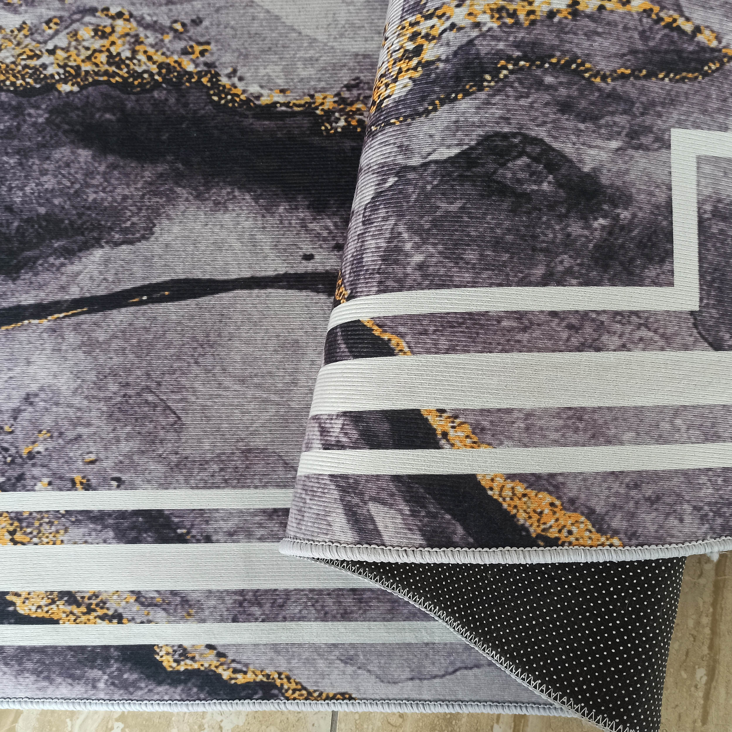Tapis aux motifs abstraits gris, noir et doré, avec des rayures blanches. Une partie est repliée, montrant le dessous antidérapant