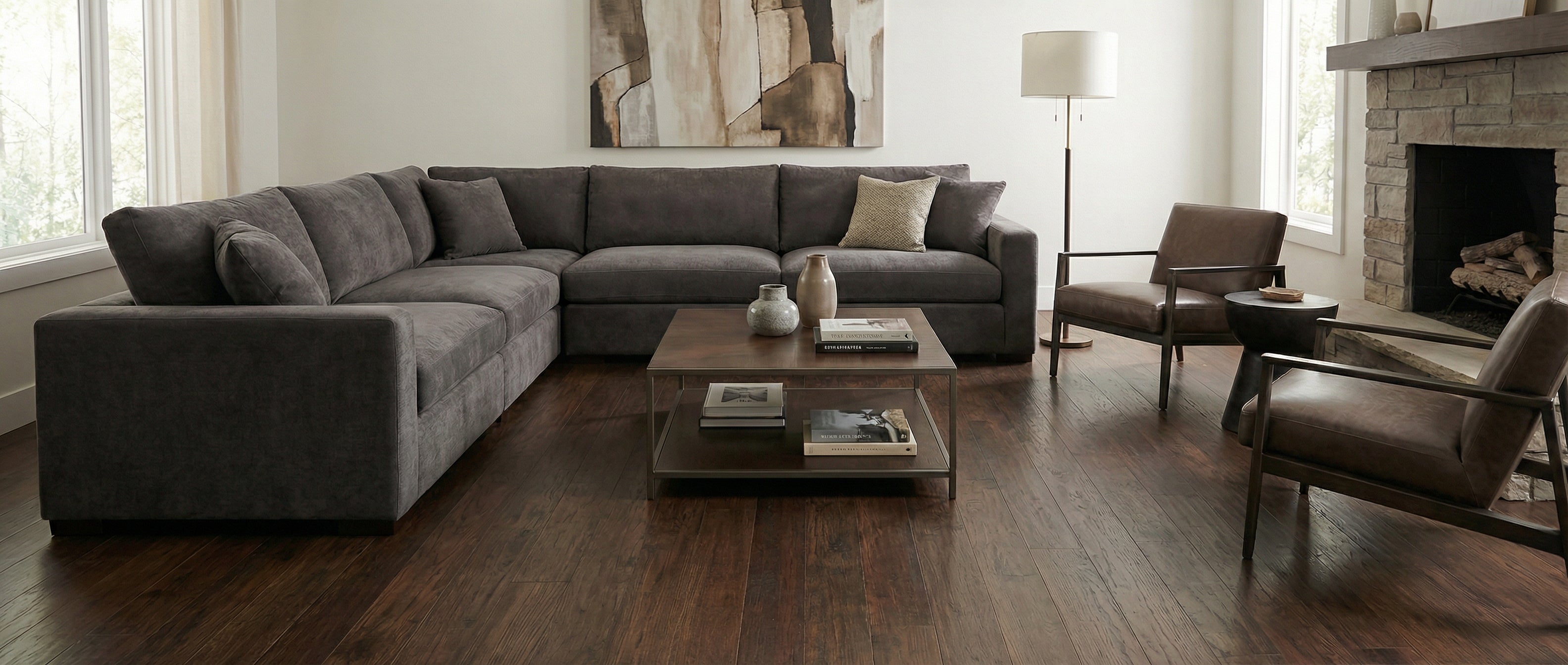 Pièce avec canapé gris, fauteuils cuir, cheminée en pierre, plancher bois foncé, table basse. Espace vide pour tapis