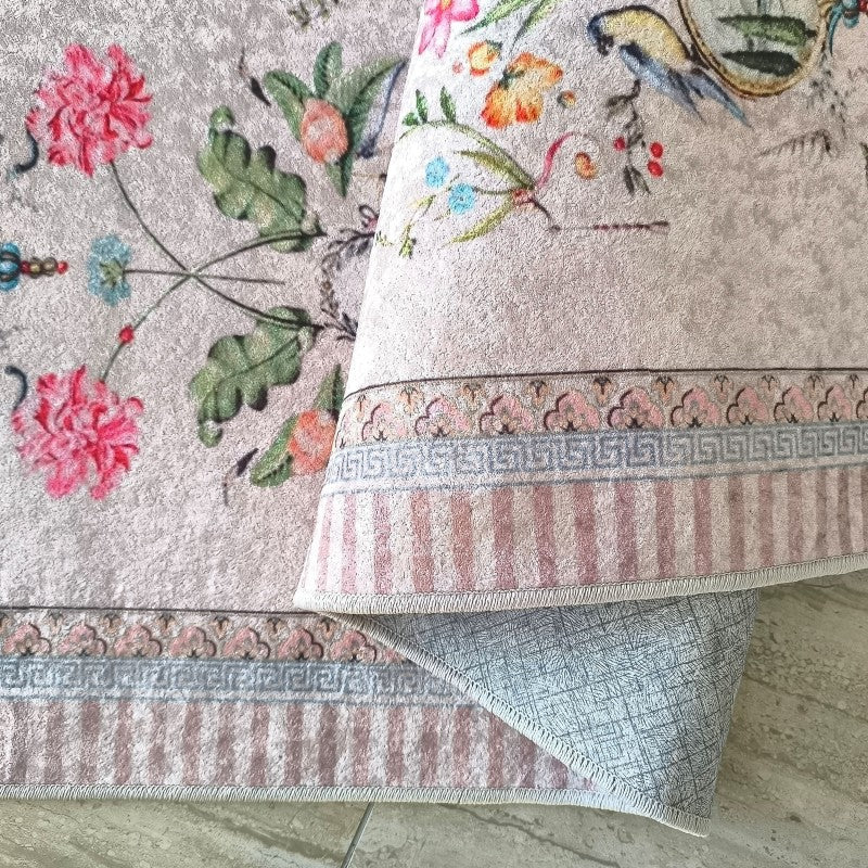 Tapis gris-rose à motifs floraux de pivoines roses et feuillages verts. Une partie est roulée, dévoilant une bordure ornementale et l'envers texturé gris