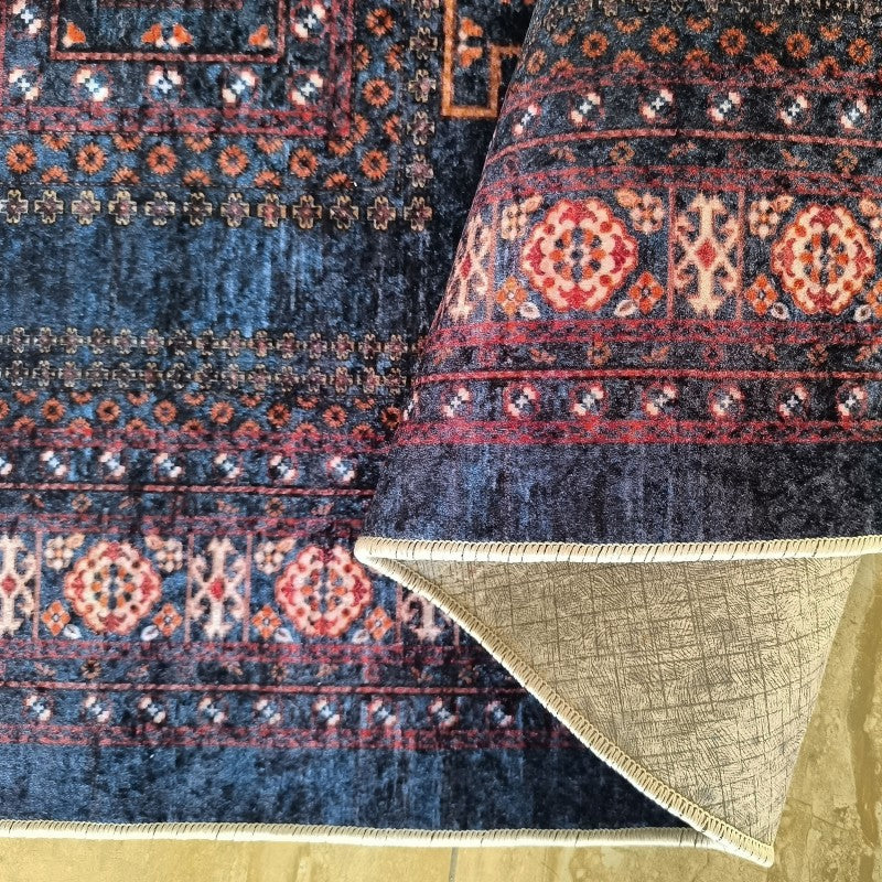 Tapis velouté bleu foncé aux motifs traditionnels rouges, oranges et beiges. Une partie est roulée, révélant son envers gris texturé