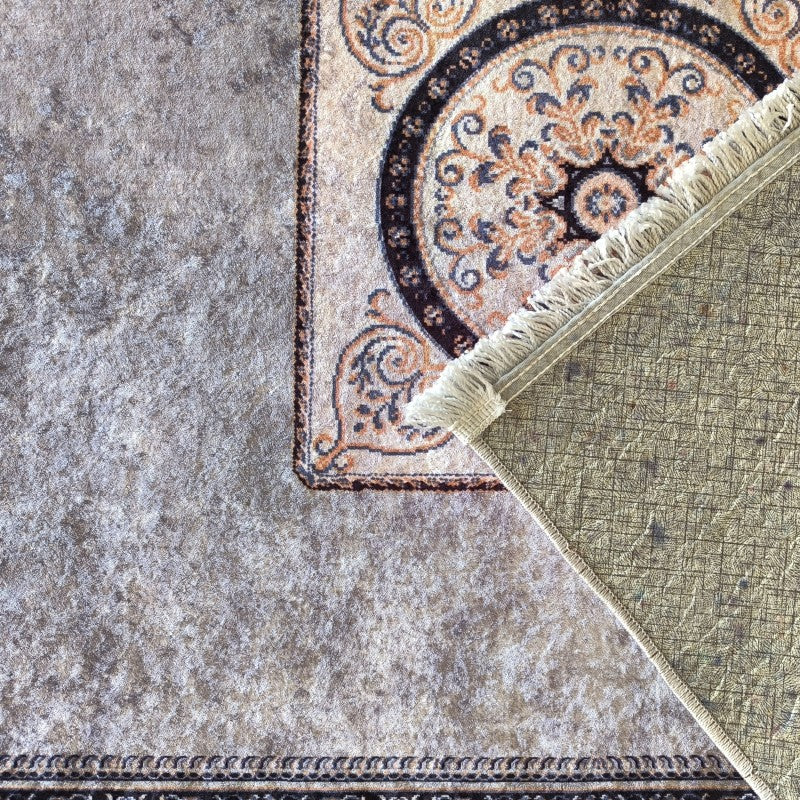 Tapis beige à motifs ornementaux marron, ocre et bleu, soulevé sur un tapis gris texturé, révélant son envers vert quadrillé et ses franges