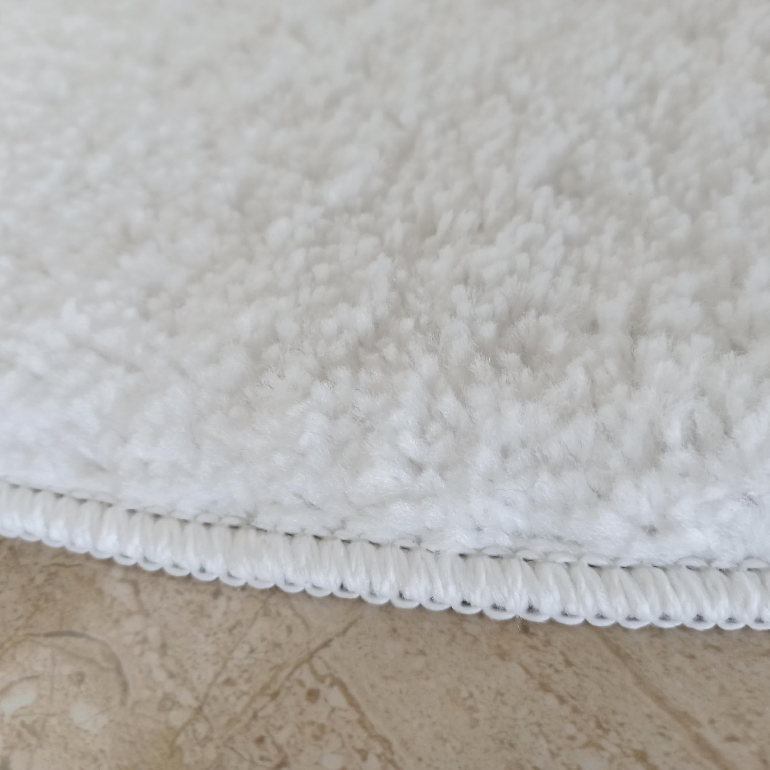 Un tapis blanc très doux et duveteux, montrant sa texture dense et son bord sur des carreaux de sol marbrés beige