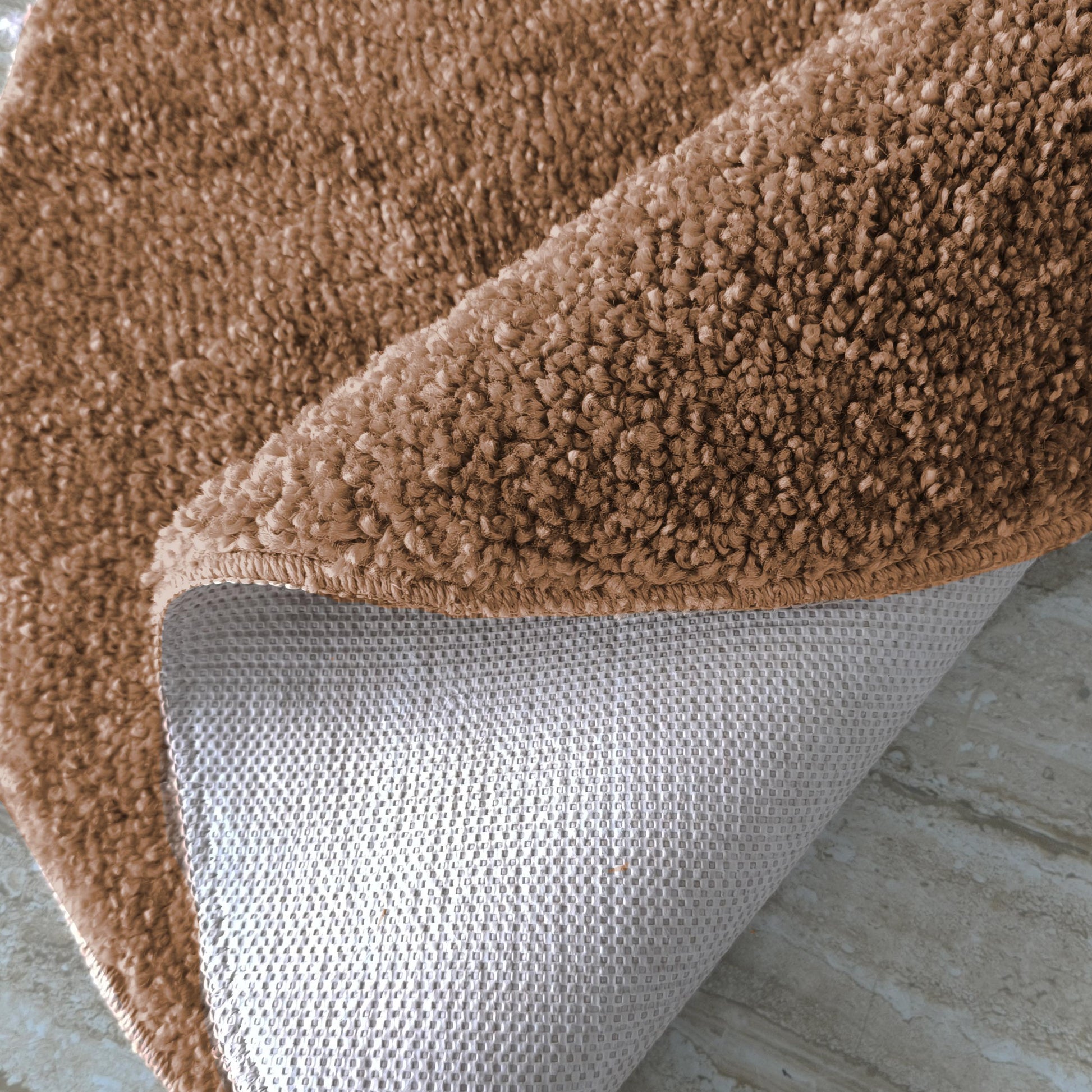Tapis marron clair à texture fourrure épaisse, soulevé pour révéler son envers gris antidérapant sur un sol pâle