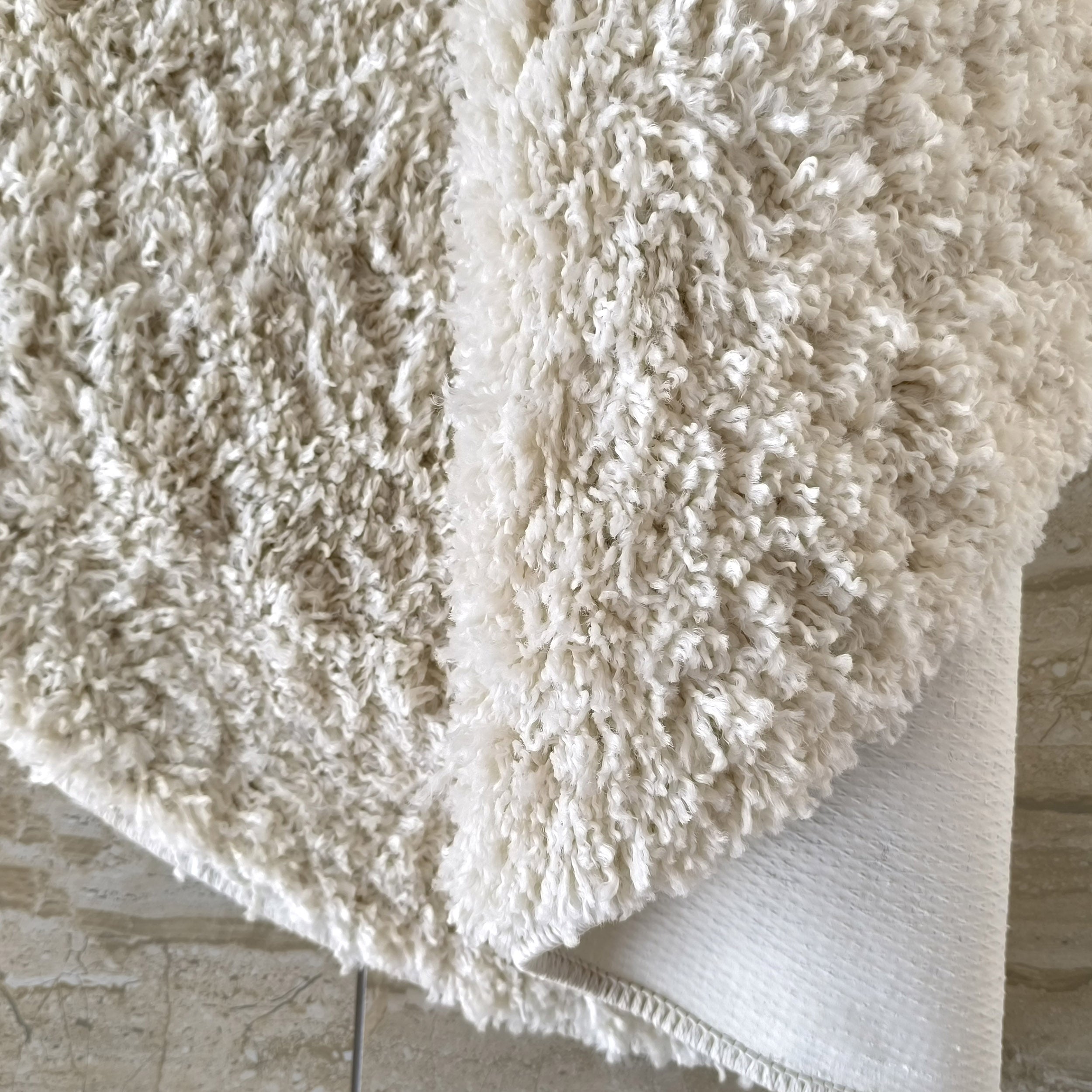 Tapis moelleux à poils longs de couleur crème et blanche, posé sur un sol carrelé beige clair, avec une partie du tapis légèrement repliée
