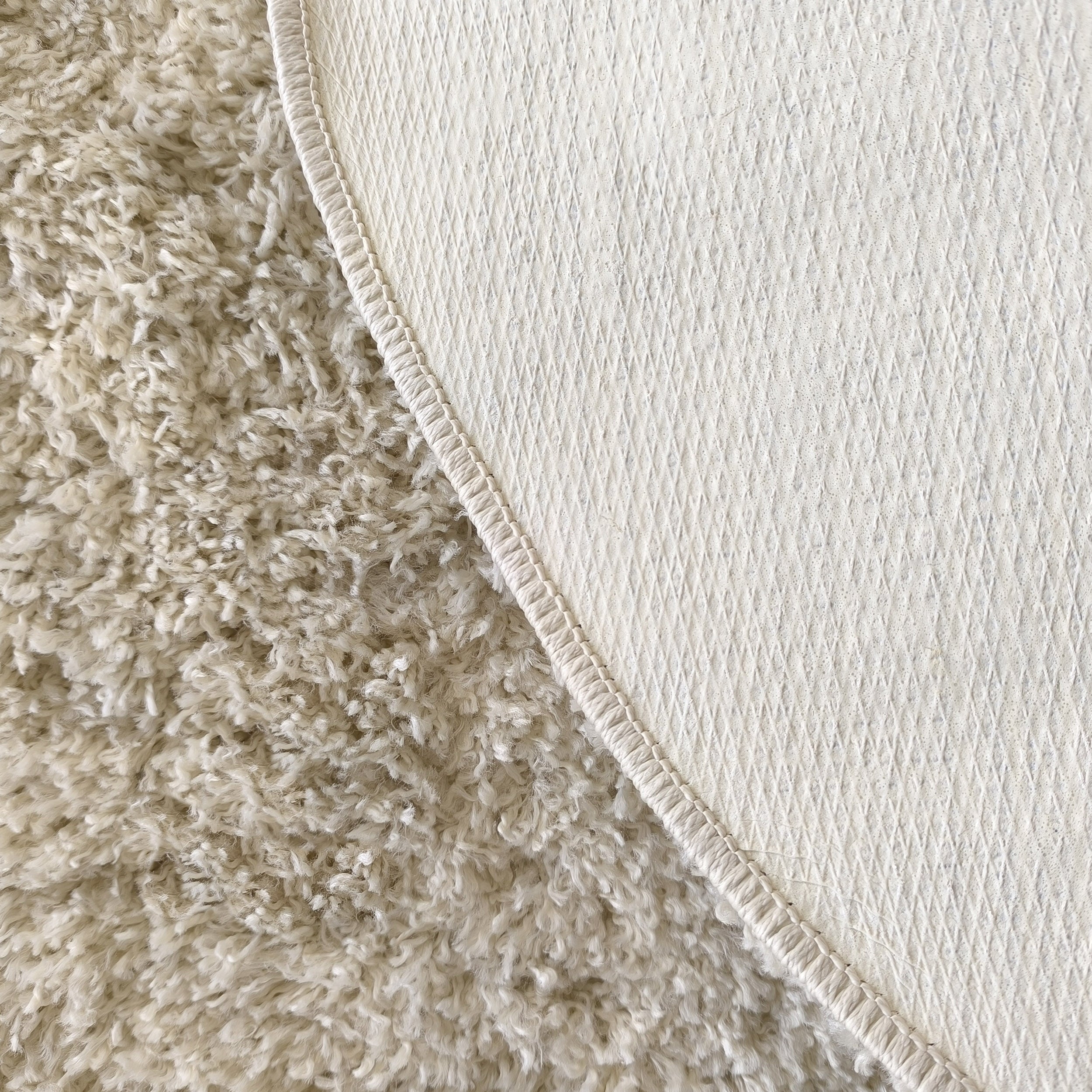 Gros plan sur un tapis beige montrant sa face supérieure moelleuse et son revers texturé à motifs losanges