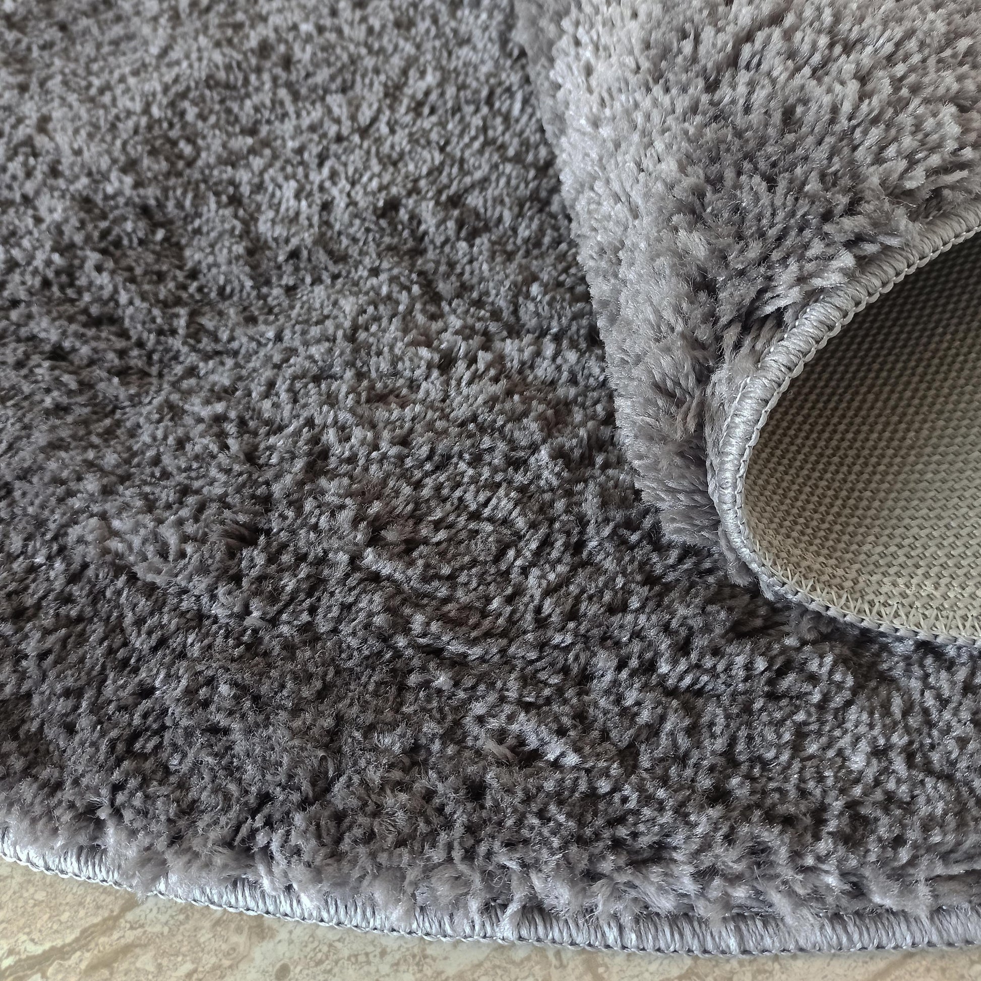 Un tapis doux gris à poils longs, dont une partie est roulée, révélant son envers tissé et les bords surpiqués