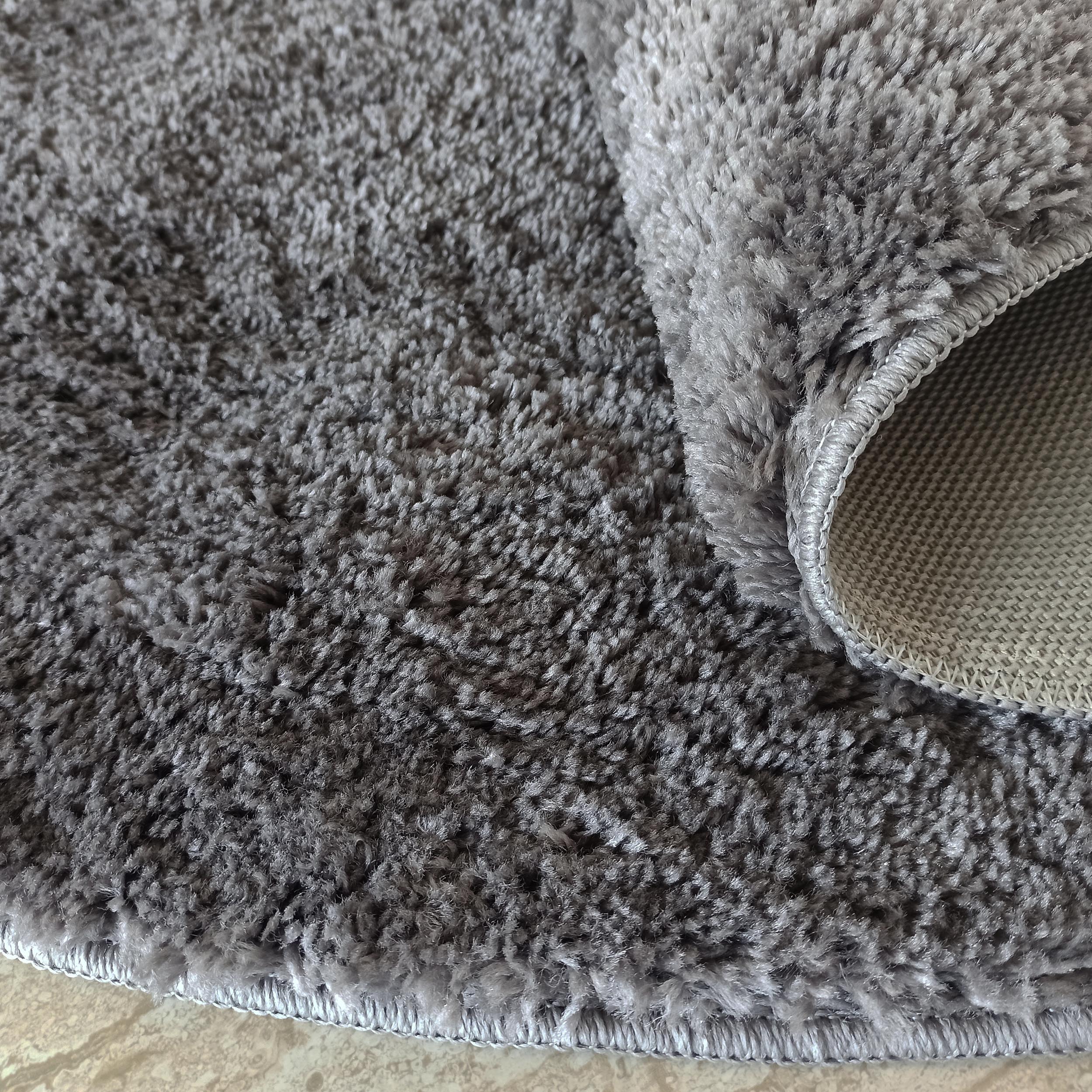 Un tapis doux gris à poils longs, dont une partie est roulée, révélant son envers tissé et les bords surpiqués