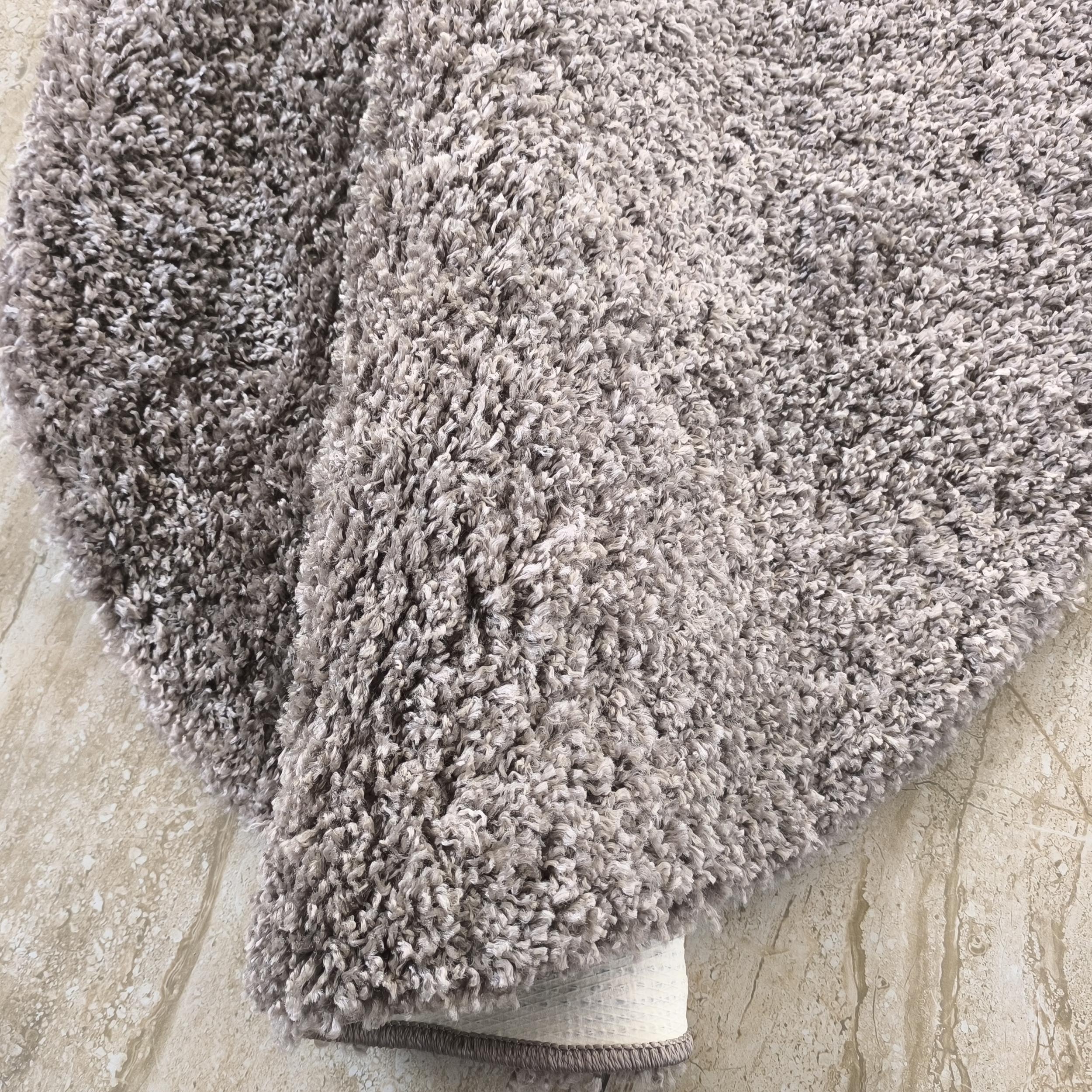 Tapis à poils longs couleur greige et blanc, partiellement enroulé sur un carrelage clair veiné, révélant sa texture duveteuse