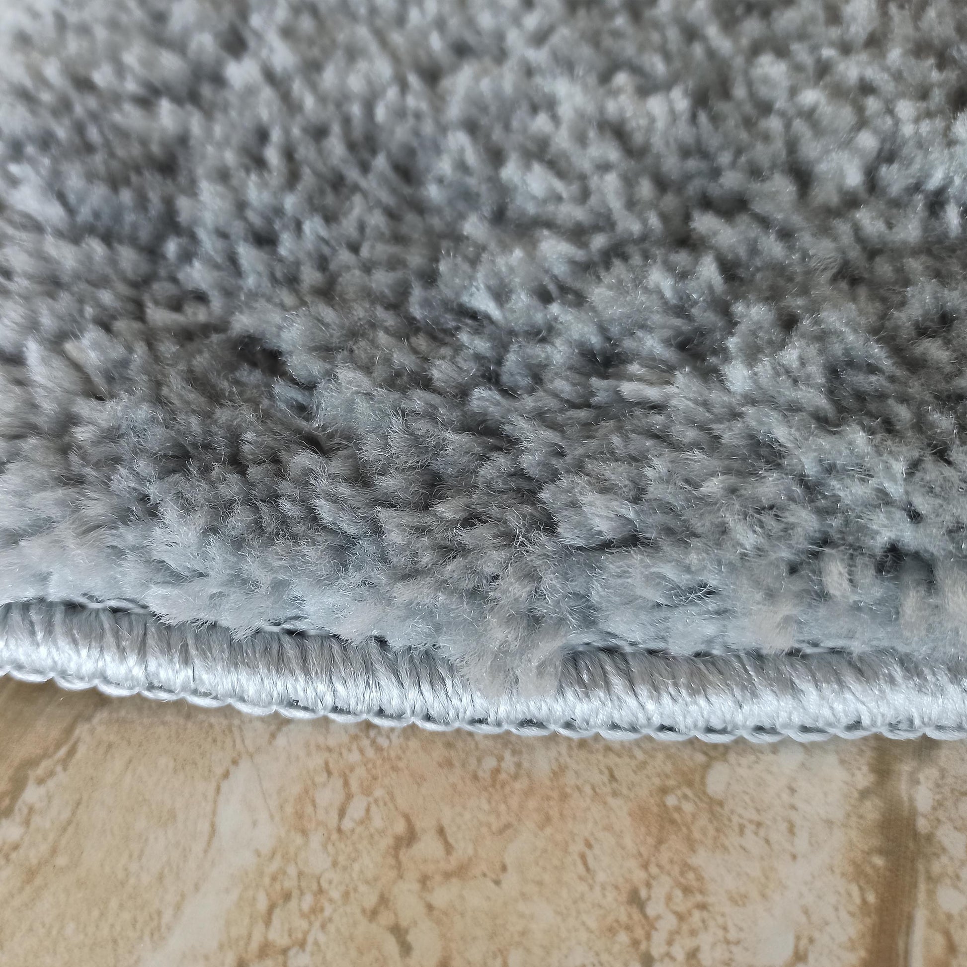 Un tapis gris clair duveteux à longs poils, avec sa bordure cousue, repose sur un sol carrelé beige marbré