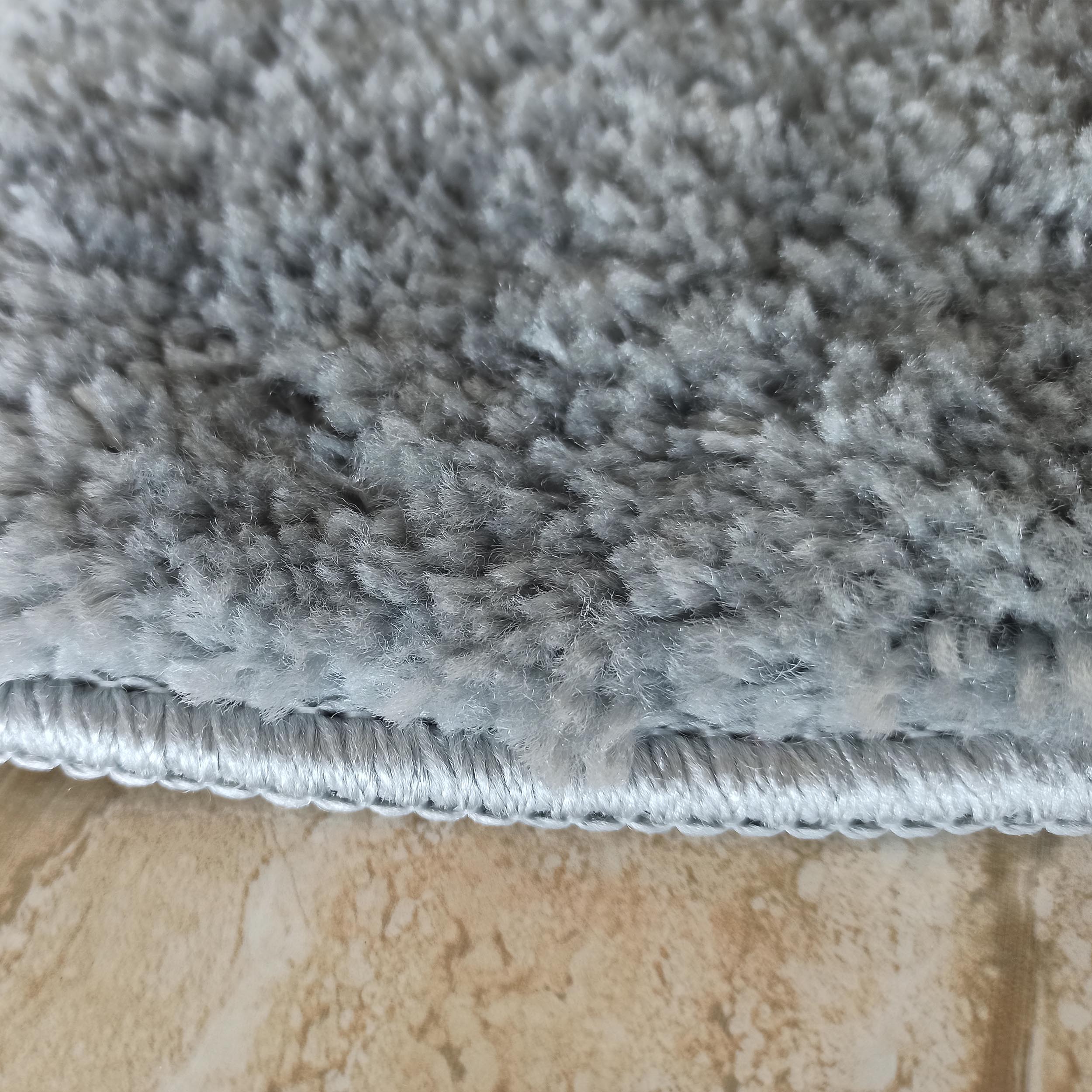Un tapis gris clair duveteux à longs poils, avec sa bordure cousue, repose sur un sol carrelé beige marbré