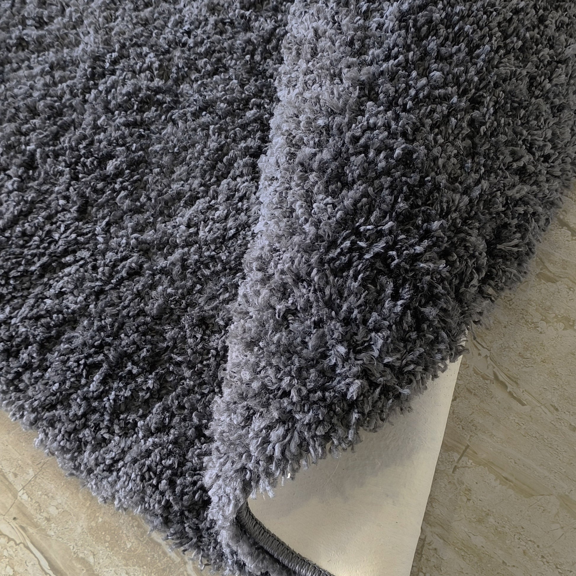 Un tapis gris foncé à longues fibres douces, légèrement enroulé sur un sol clair, dévoilant son envers beige