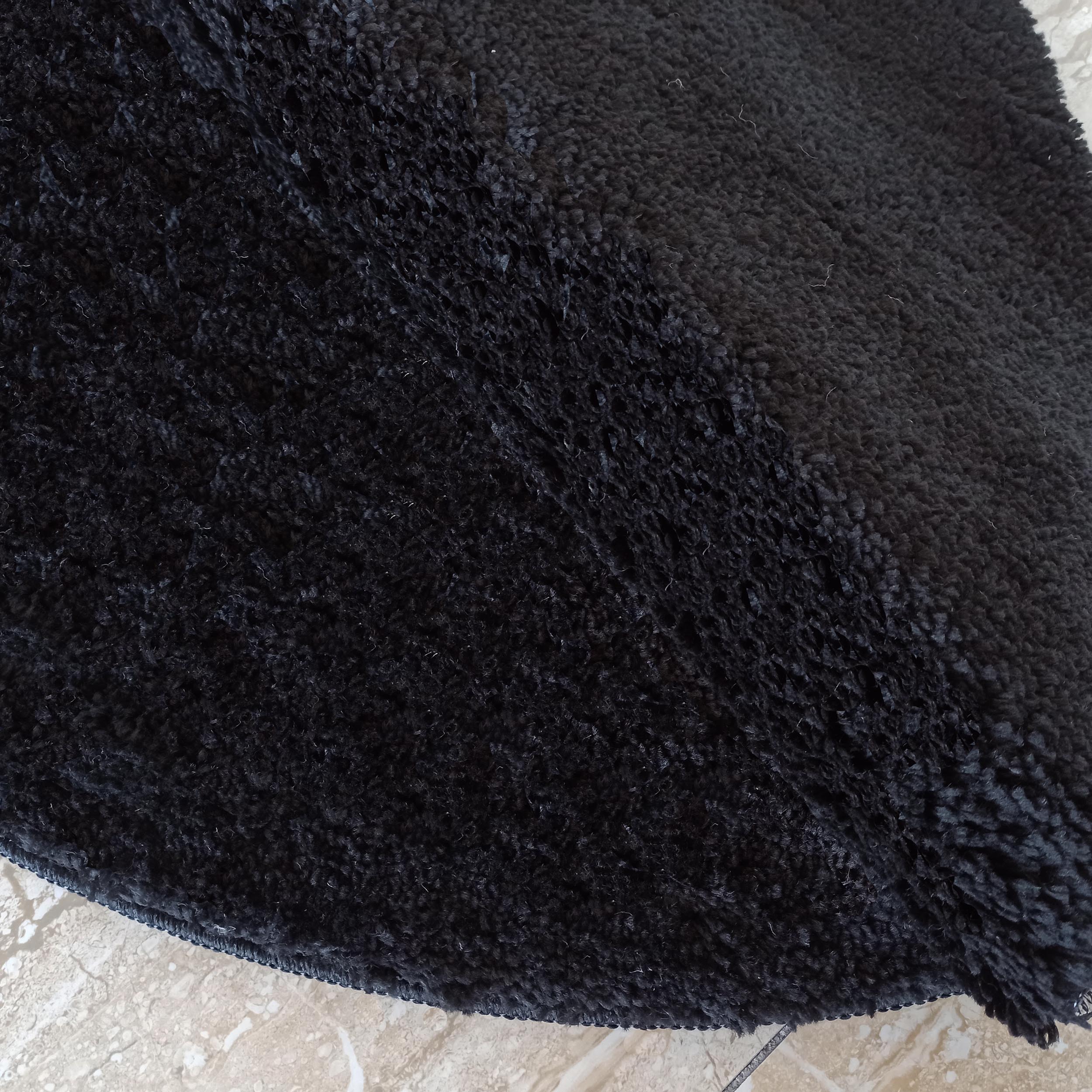 Tapis noir à poils longs et texture shaggy, posé sur un sol clair marbré. La surface est dense et moelleuse