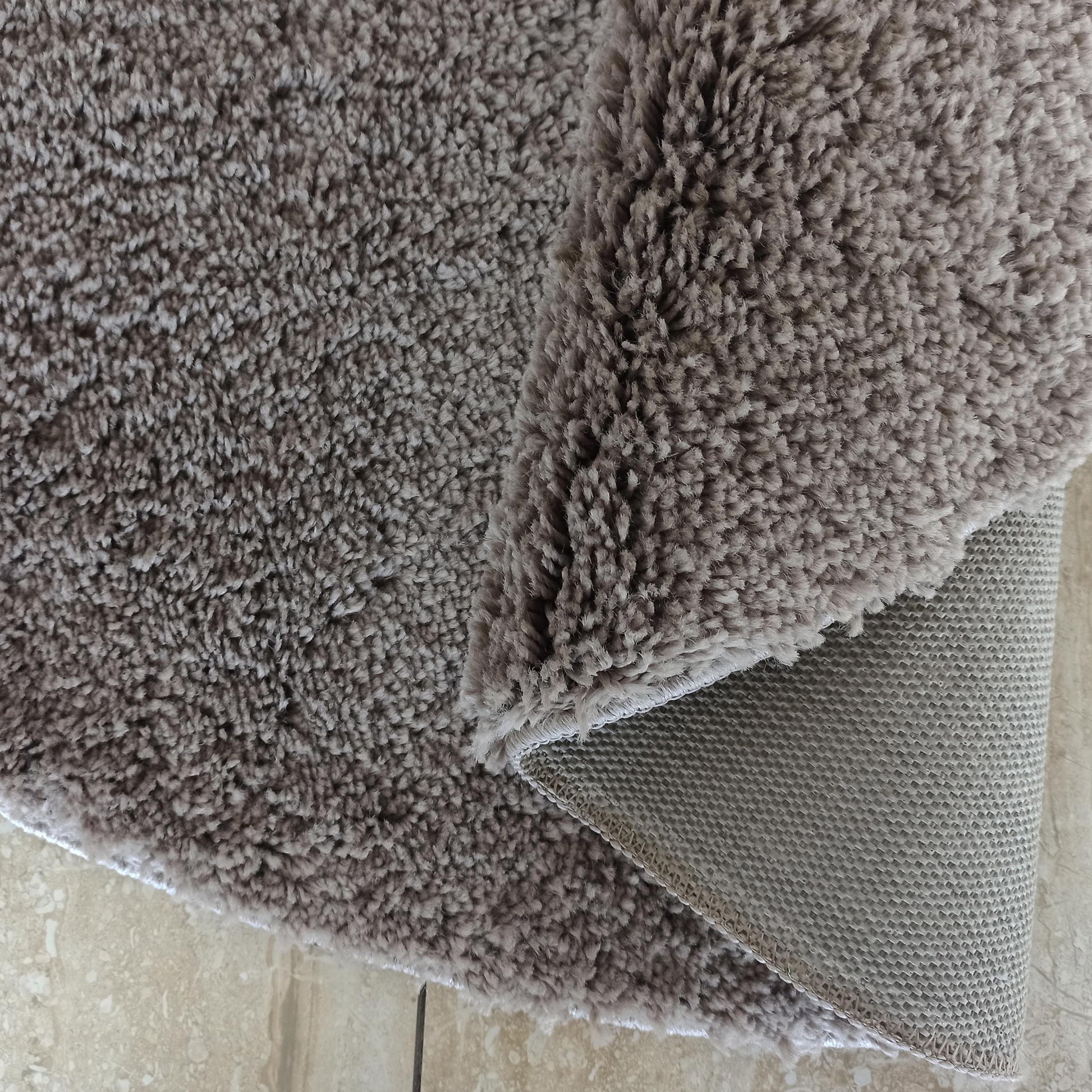 Tapis doux à poils longs gris-taupe, une section est repliée révélant son envers texturé sur un sol clair