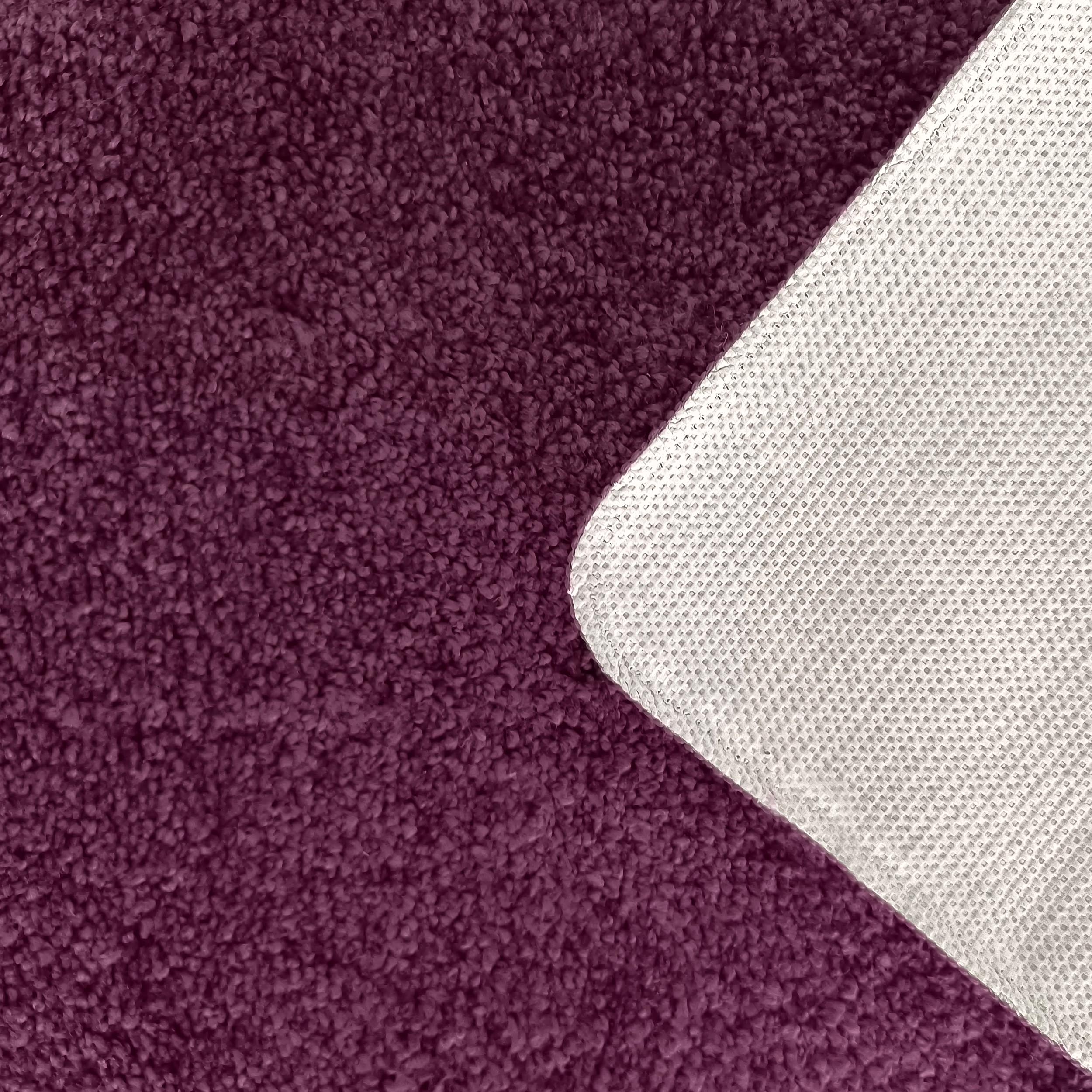 Gros plan sur un tapis moelleux de couleur aubergine, avec un coin relevé montrant son envers texturé gris clair à motif quadrillé